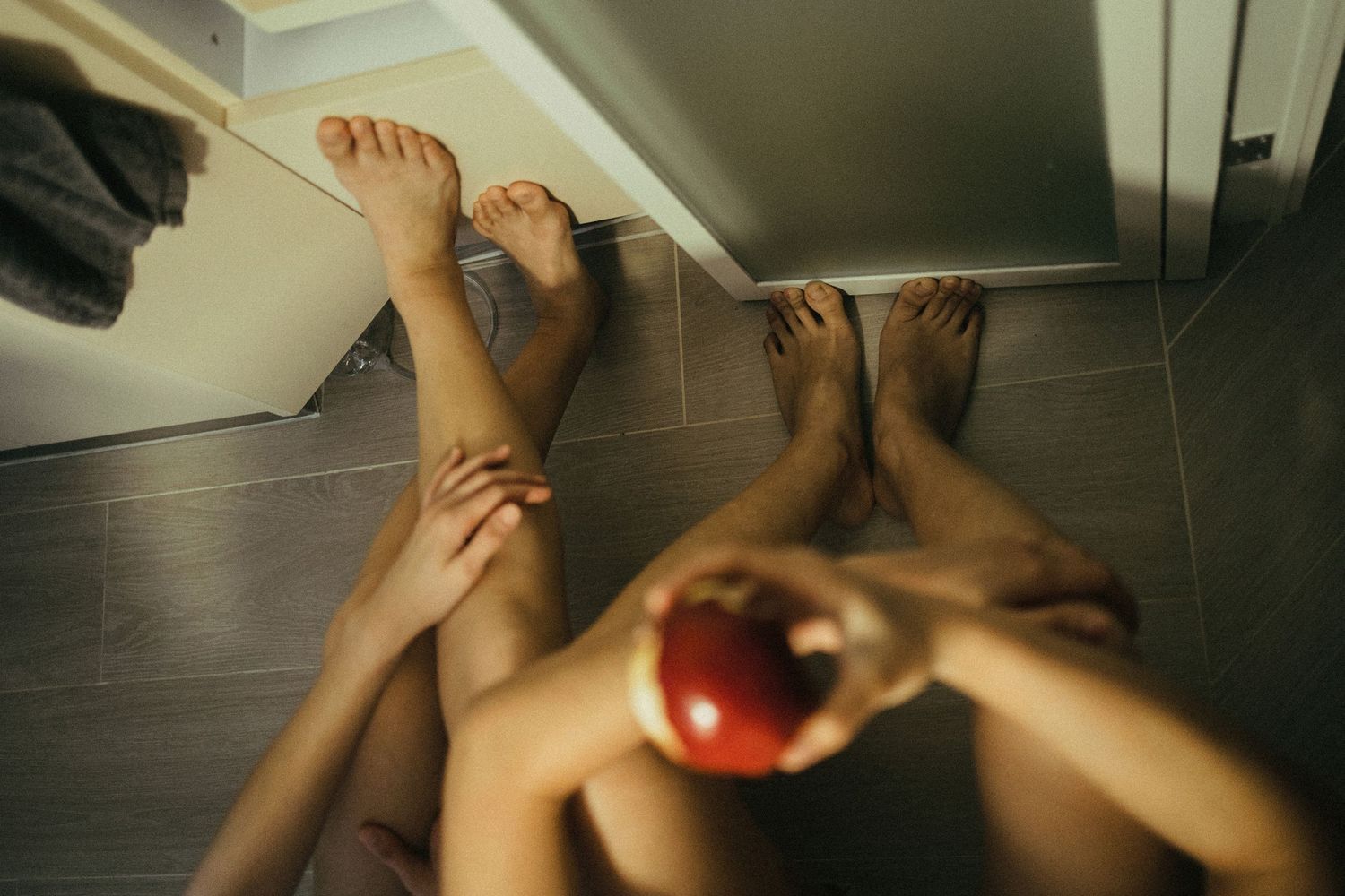 Two people sitting on a bathroom floor, one holding a red apple, their bare legs intertwined on gray tiles.
