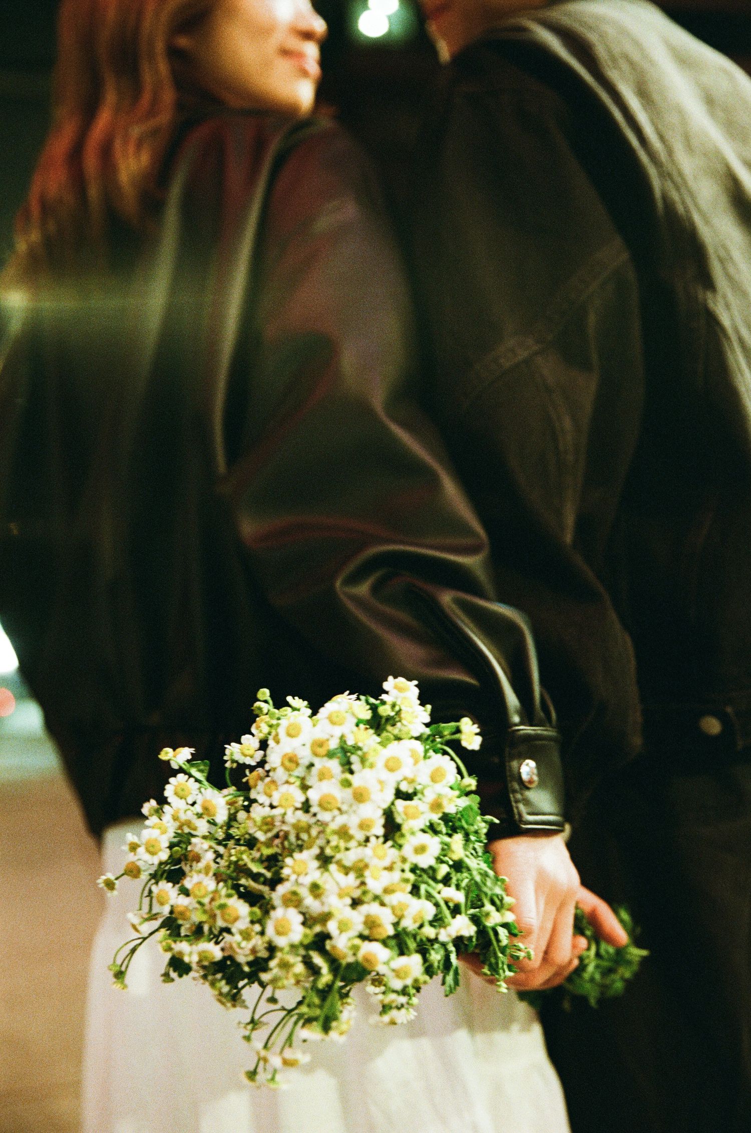 Couple standing close together at night, holding a bouquet of white daisy flowers.