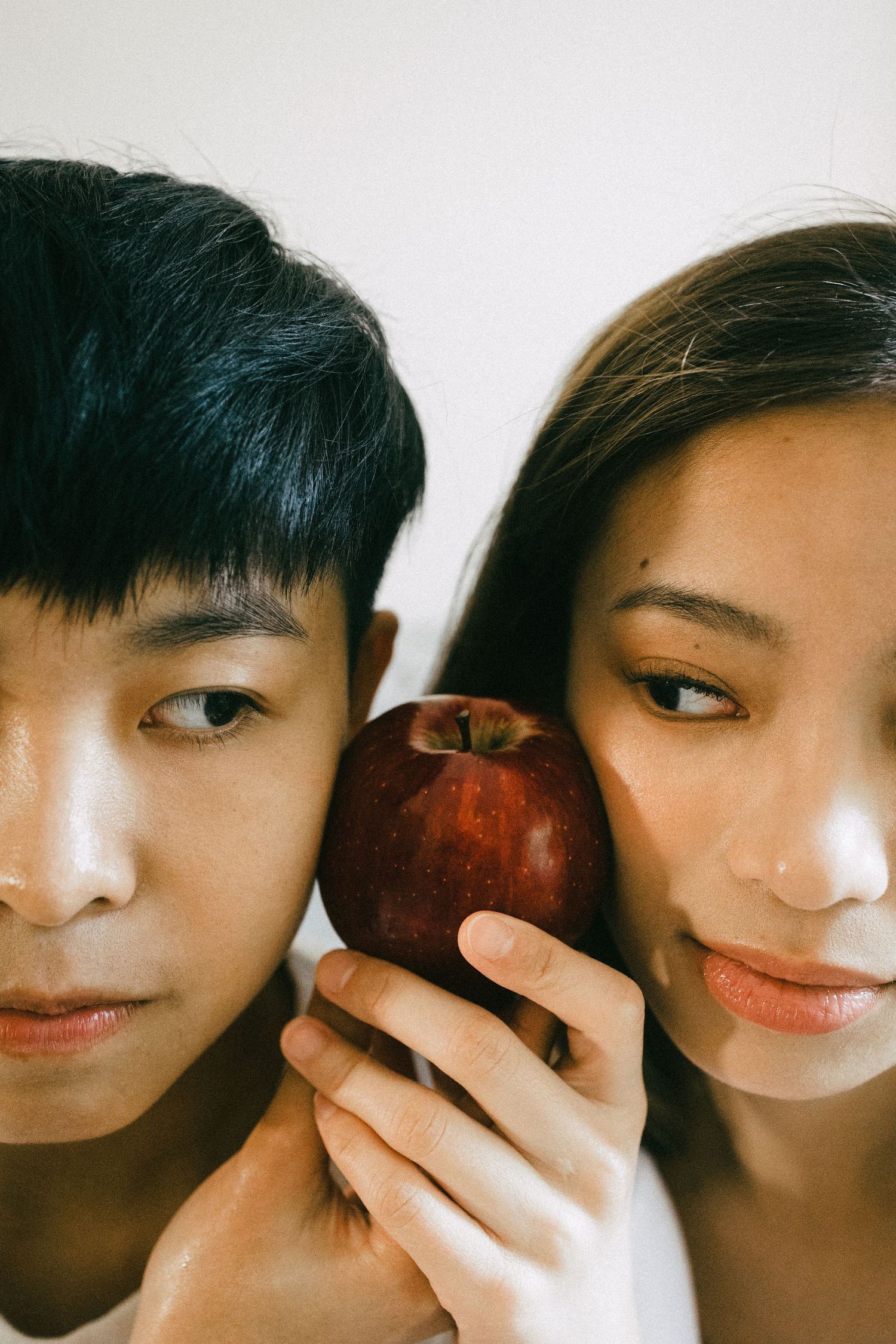 A close-up portrait of a young Asian couple holding a red apple between their faces.