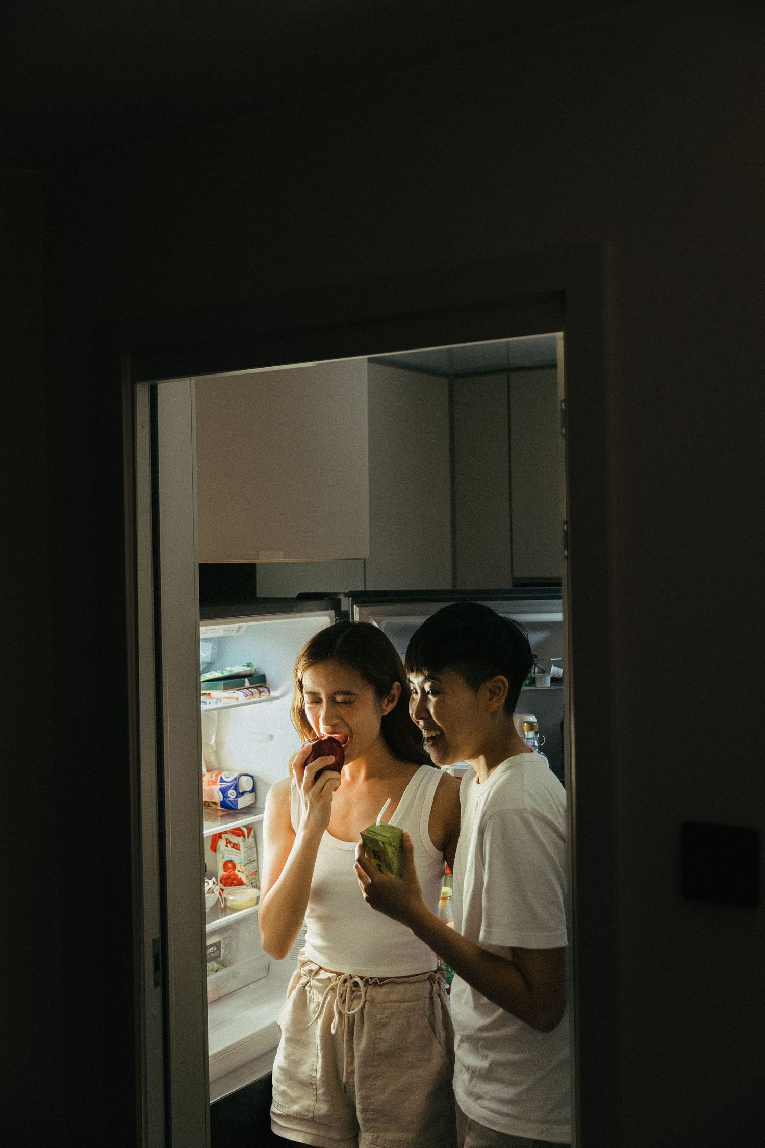 A couple standing by an open refrigerator at night, woman eating a red apple in a dark kitchen.