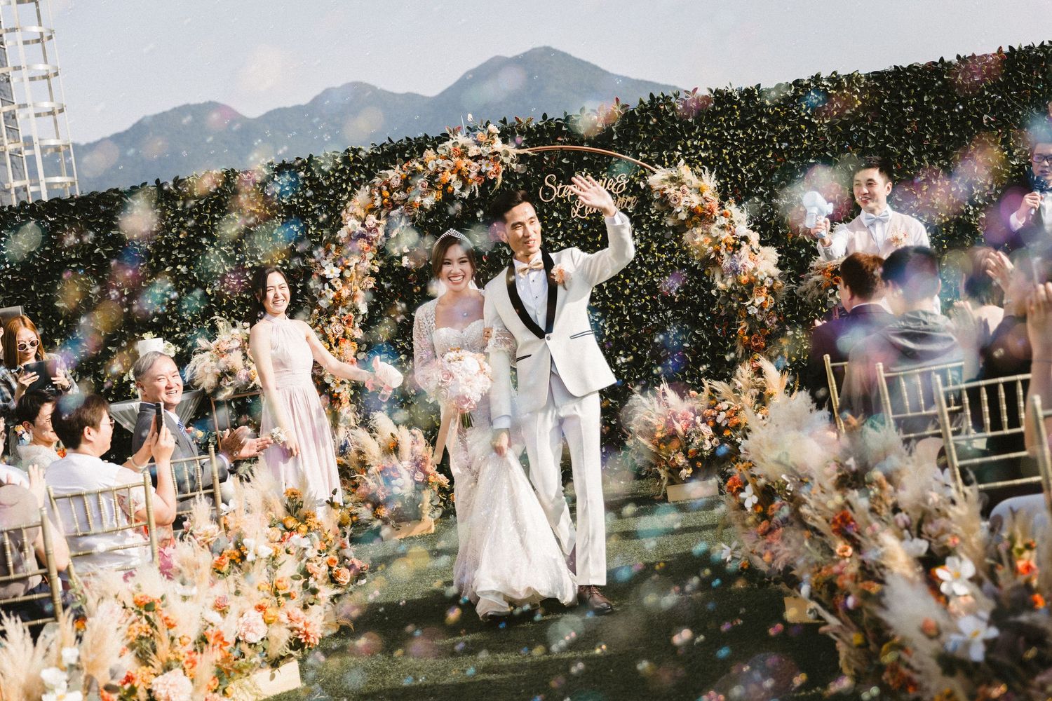 A bride and groom exit through a floral arch with confetti flying at an outdoor mountain wedding.