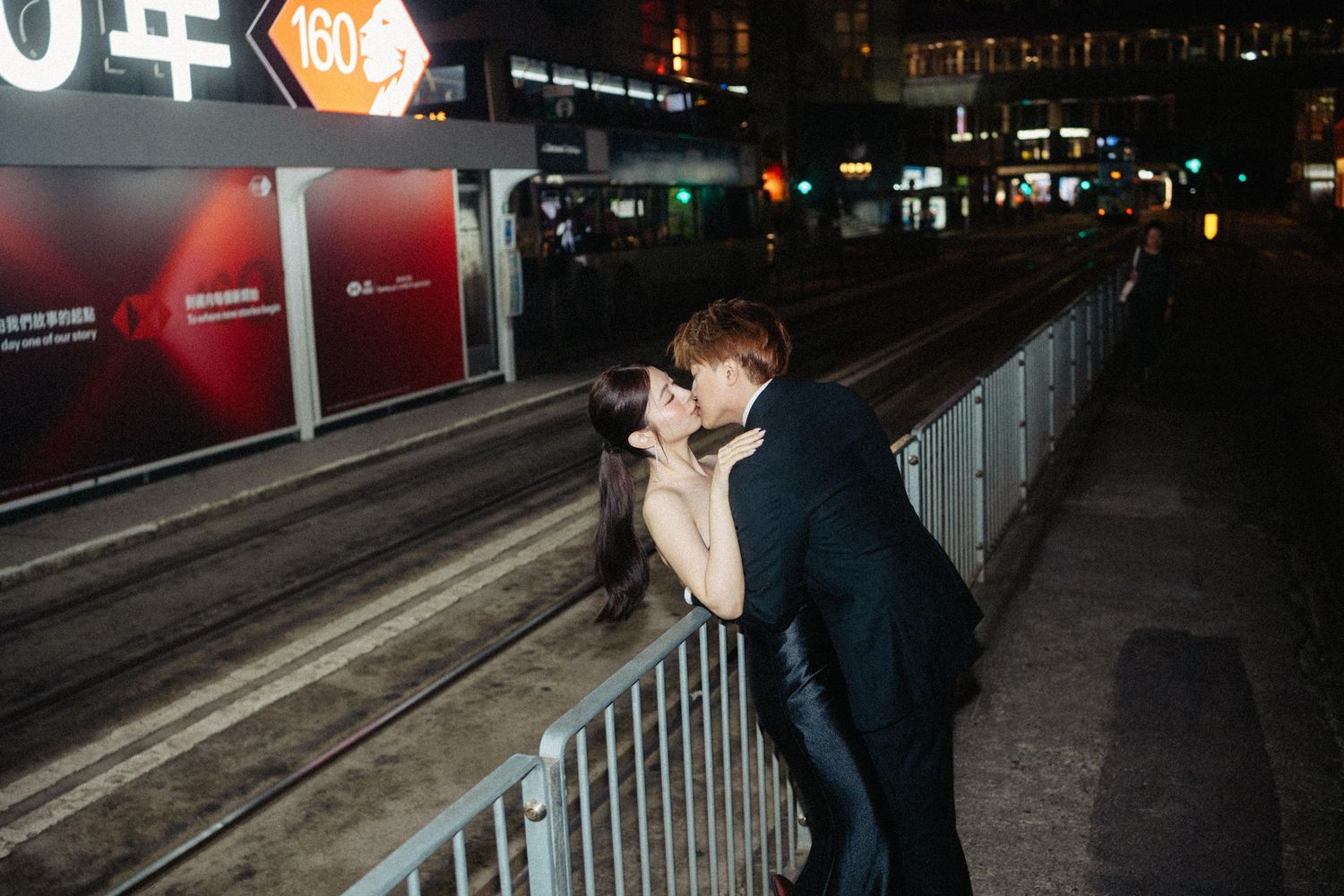 A couple in elegant evening wear shares a passionate kiss at night beside tram tracks with city lights behind them.