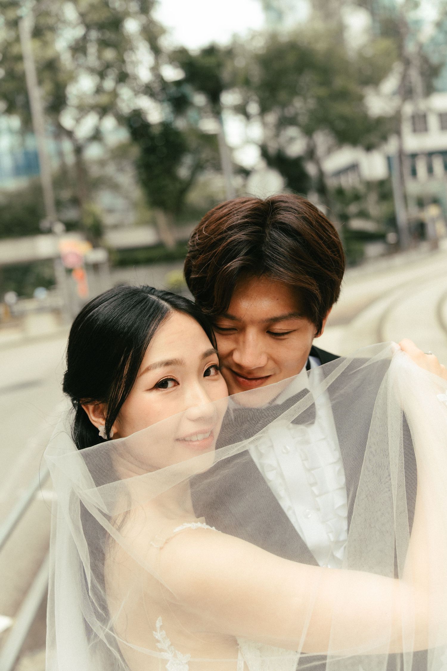 A smiling Asian bride and groom share a tender moment beneath a flowing white veil outdoors.