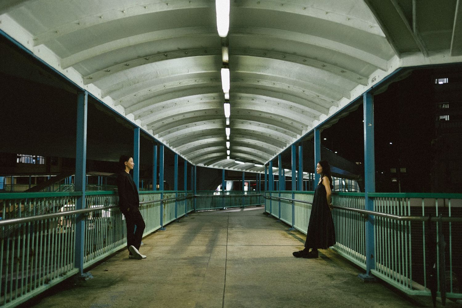 Two people standing apart on a covered pedestrian bridge at night with blue railings.