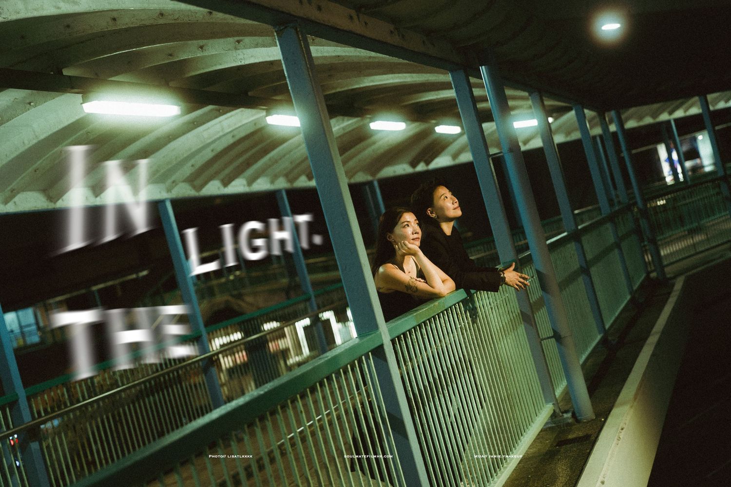 Couple standing at railing on elevated walkway at night with illuminated In The Light signage.
