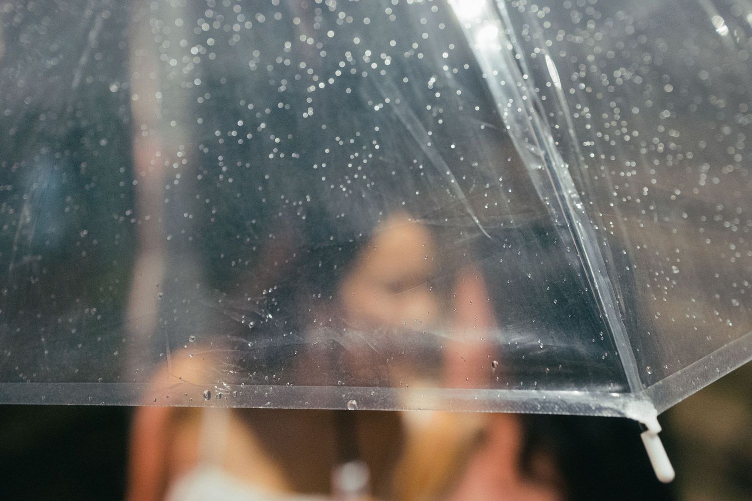 Two people share an intimate moment beneath a clear rain-dotted transparent umbrella.
