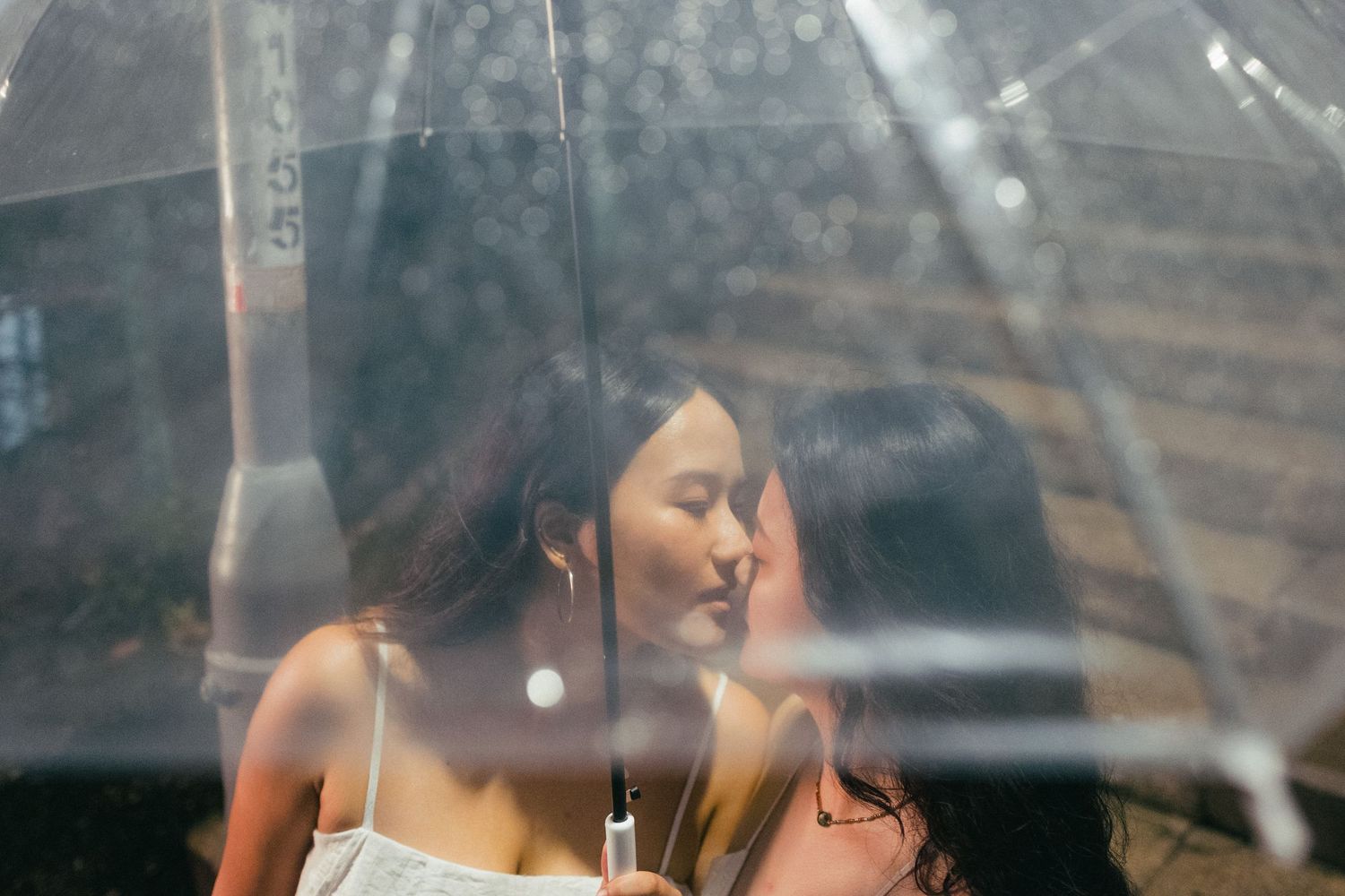 Two women sharing an intimate moment under a clear umbrella in the rain.