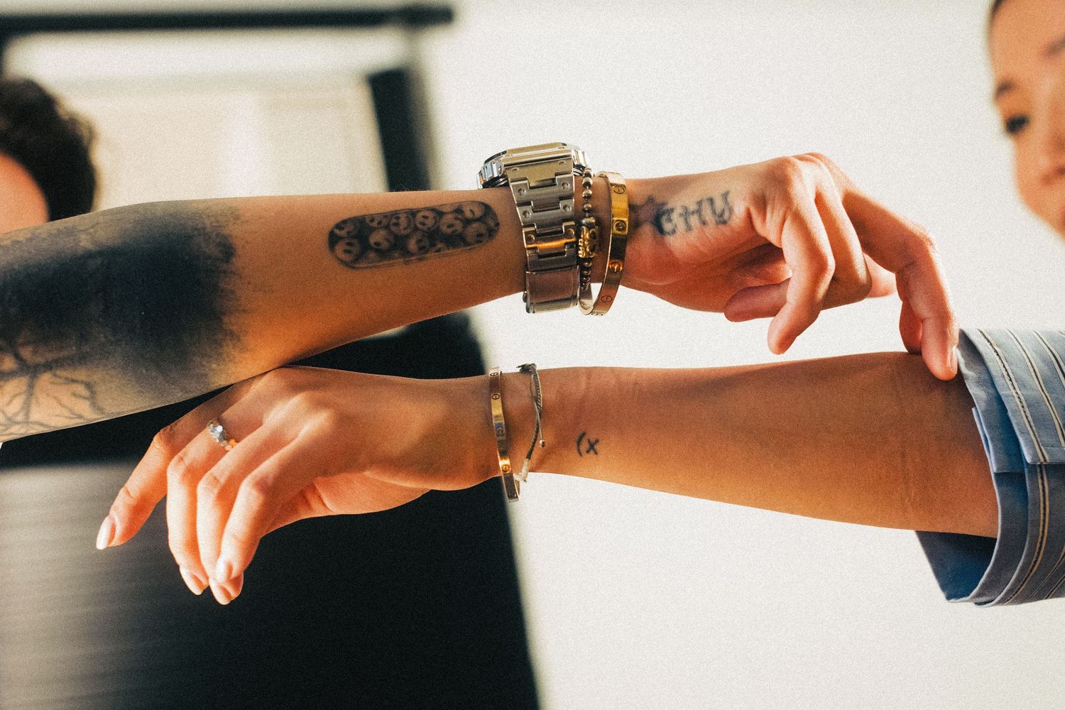 Two people comparing tattoos and bracelets on their outstretched arms indoors.