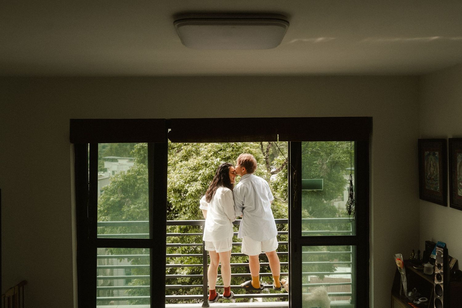 Couple in white robes standing together at glass balcony doors overlooking lush green trees.