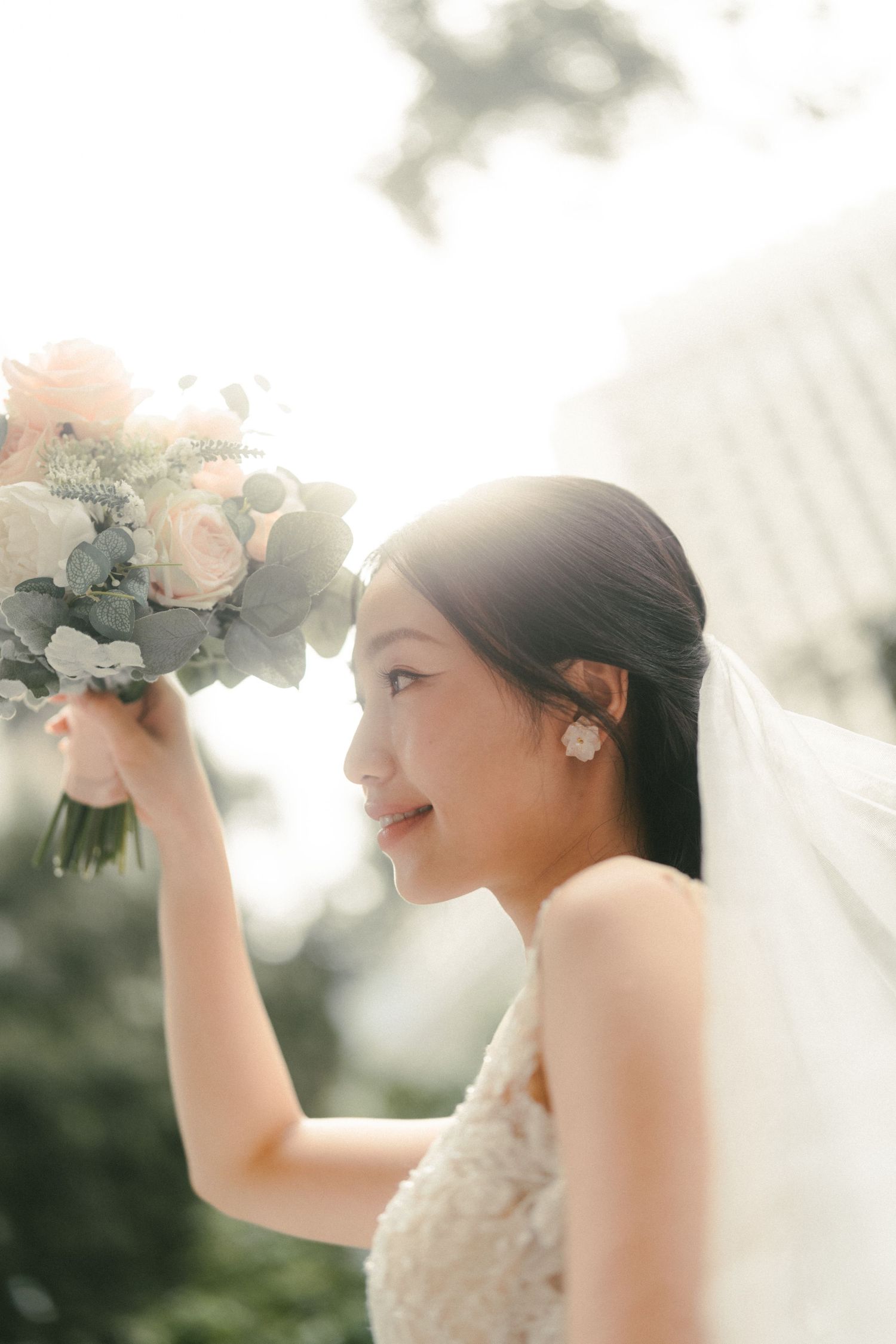 A radiant Asian bride in a lace gown holds a blush rose bouquet, smiling softly in warm backlit sunlight.