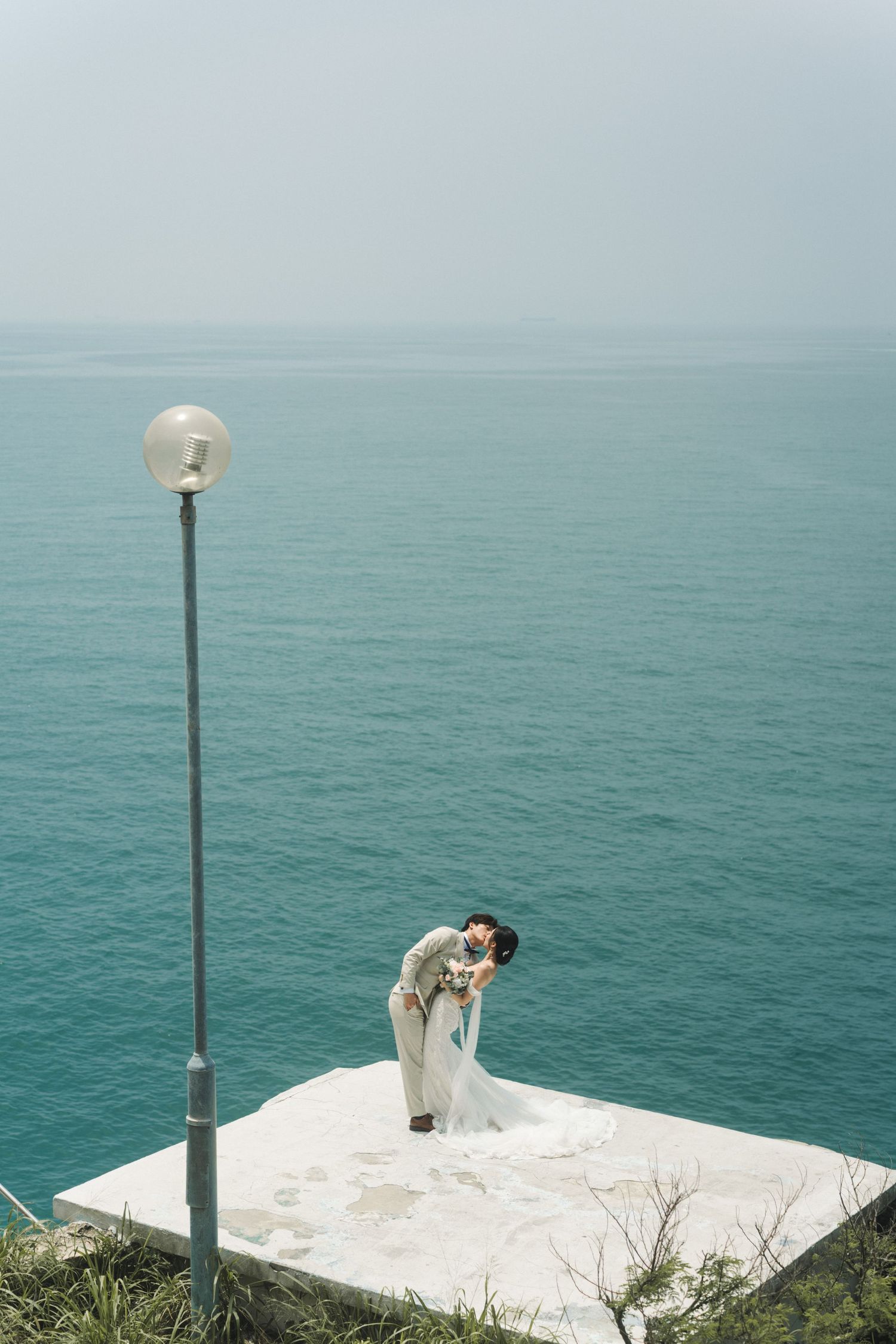 Wedding couple share a romantic dip kiss on coastal platform overlooking turquoise sea with lamp post.