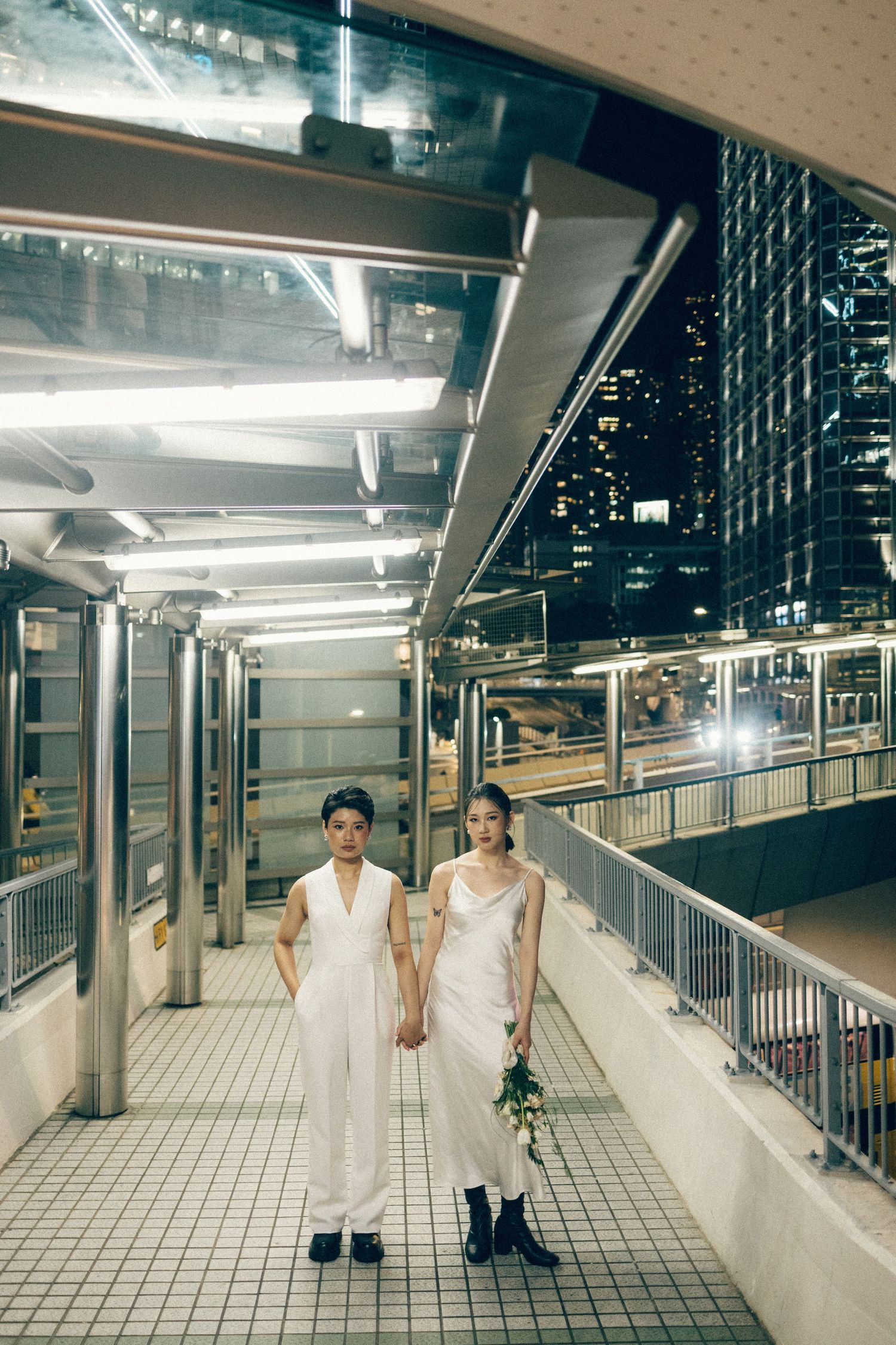 Two women in white outfits hold hands on a urban pedestrian bridge at night with city lights glowing behind them.