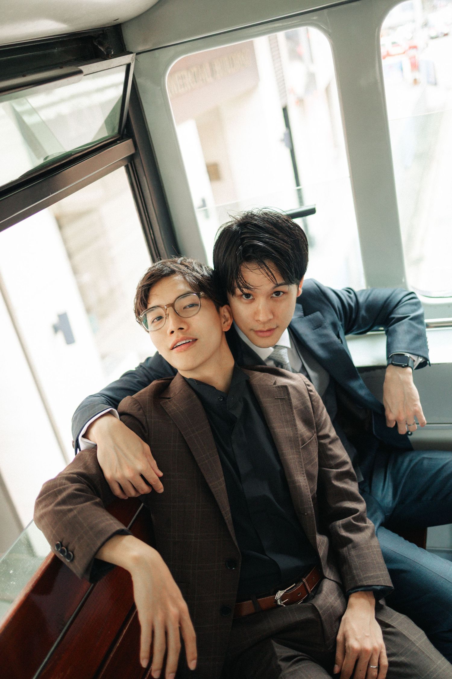 Two well-dressed Asian men sit together inside a vintage tram, one resting on the others shoulder.