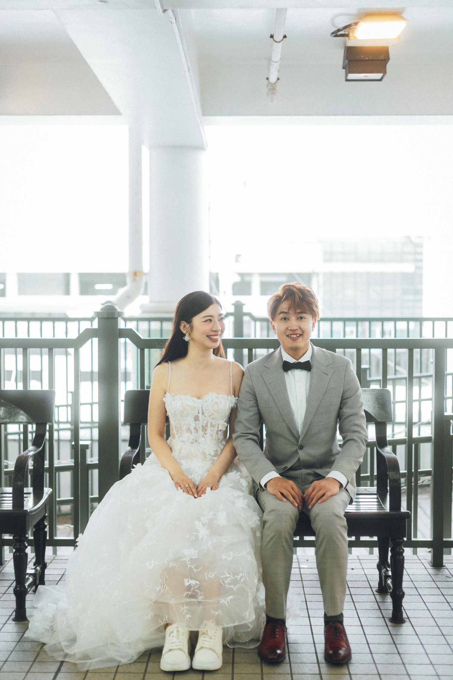 A cheerful wedding couple sits side by side in a bright ferry terminal, bride in white gown and groom in gray suit with bow tie.