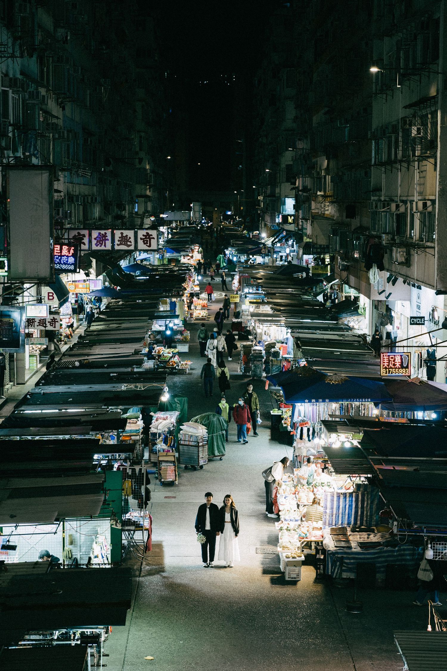 Aerial night view of a bustling Hong Kong street market with glowing neon signs and vendors lining both sides.