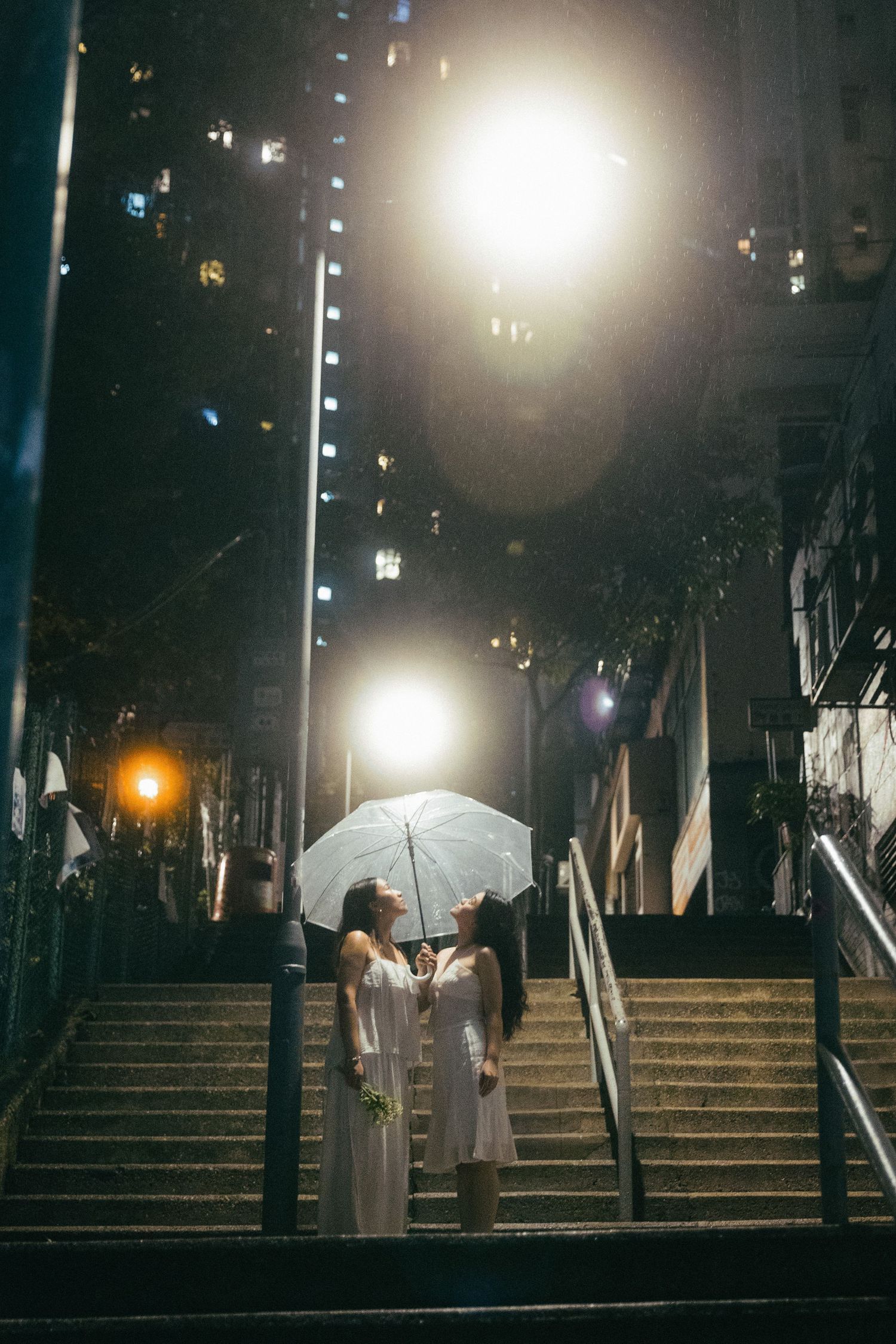 Two women in white dresses sharing a clear umbrella on rain-soaked city stairs at night.