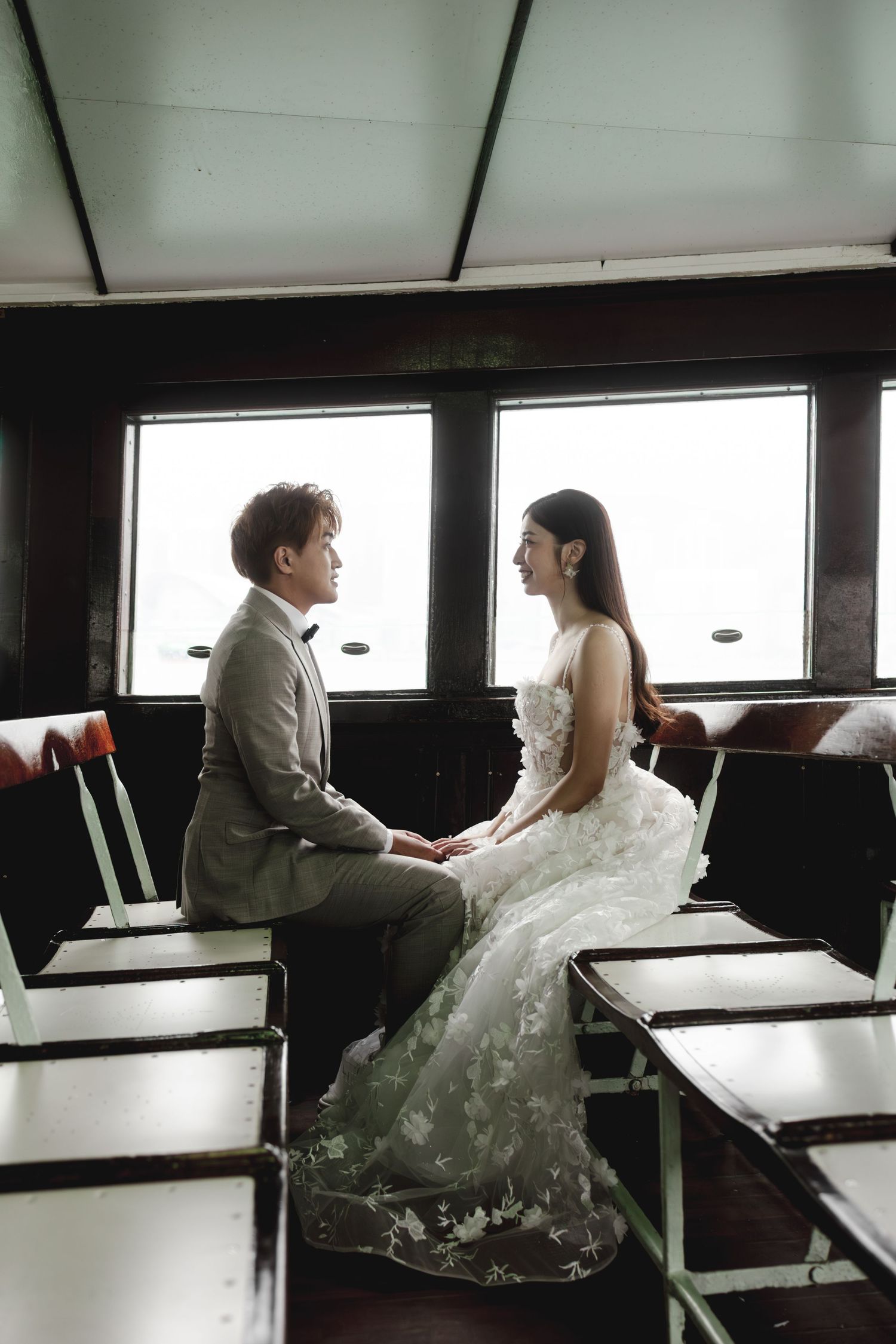 Bride and groom sit facing each other holding hands inside a vintage ferry boat interior.