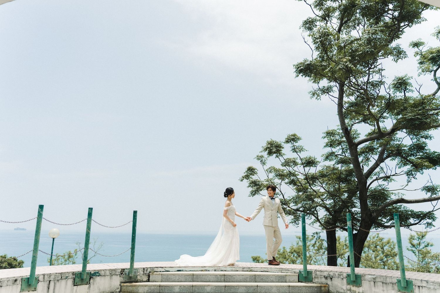 Bride and groom hold hands on scenic ocean overlook stairs during outdoor wedding photo session.