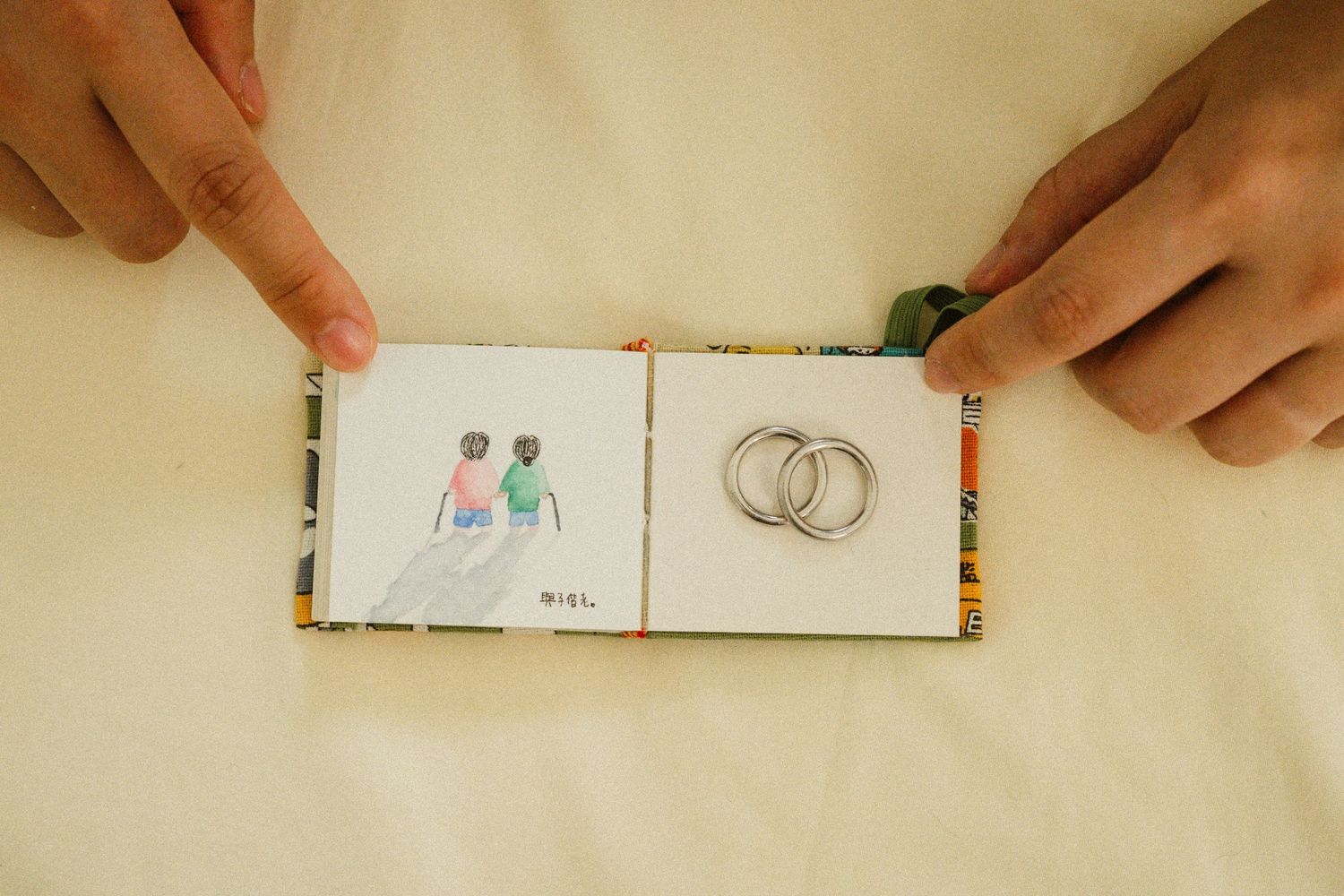 Hands open a small illustrated book beside two wedding rings on a cream background.