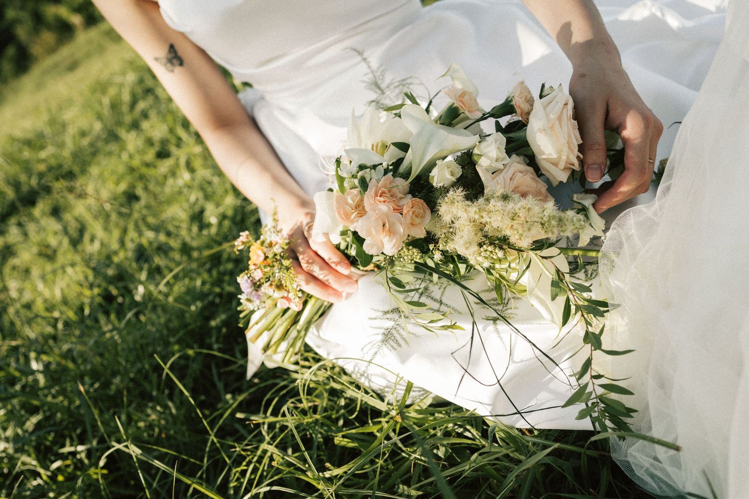 Bride holding an elegant white and blush wedding bouquet with greenery while seated in grass.