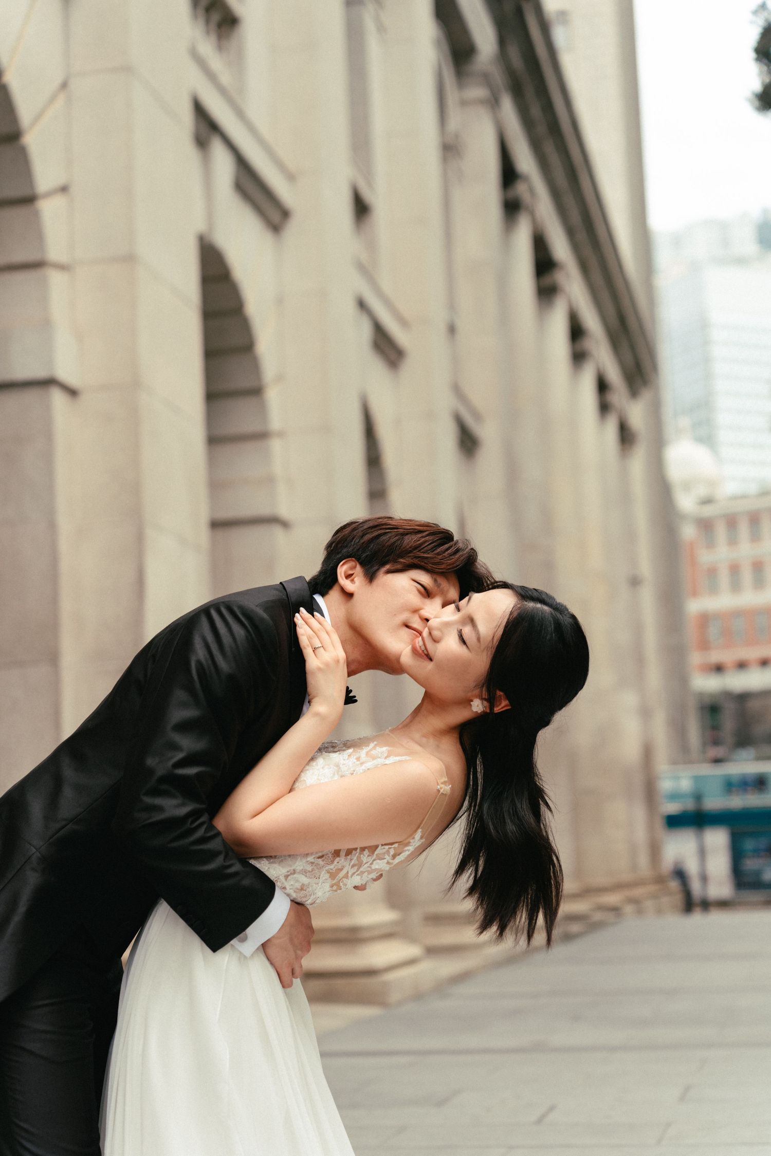 A groom dips his bride for a kiss beside a grand stone building, her white gown flowing elegantly.
