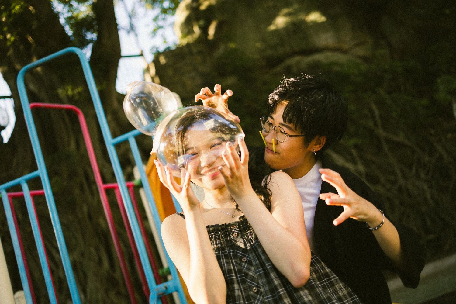 Playful couple laughing outdoors as woman holds a large soap bubble in front of her face.