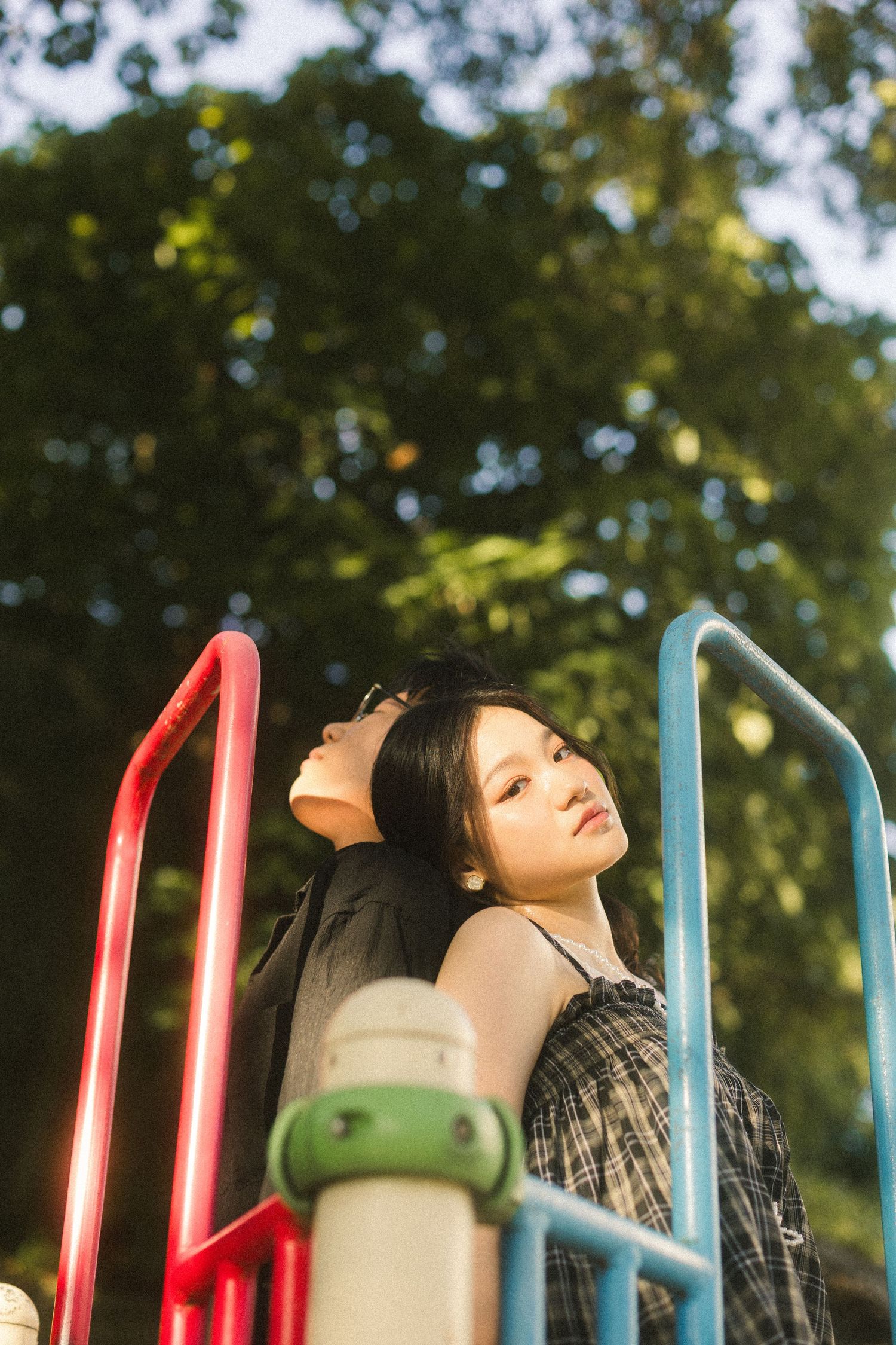 Young woman leaning back on colorful playground equipment surrounded by lush green trees in warm sunlight.