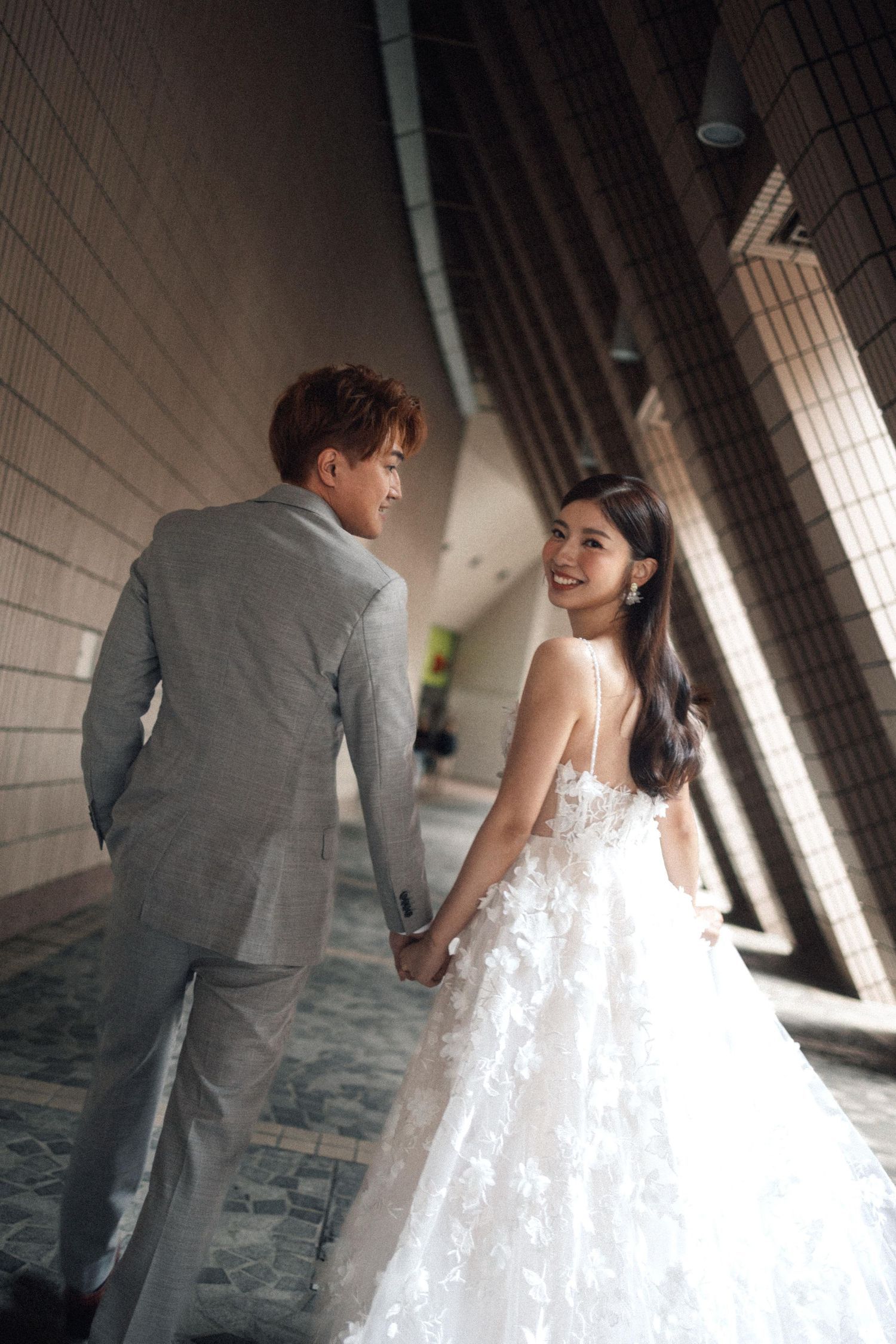 A bride in a floral white gown smiles back at her groom in a gray suit as they walk hand in hand through a modern corridor.