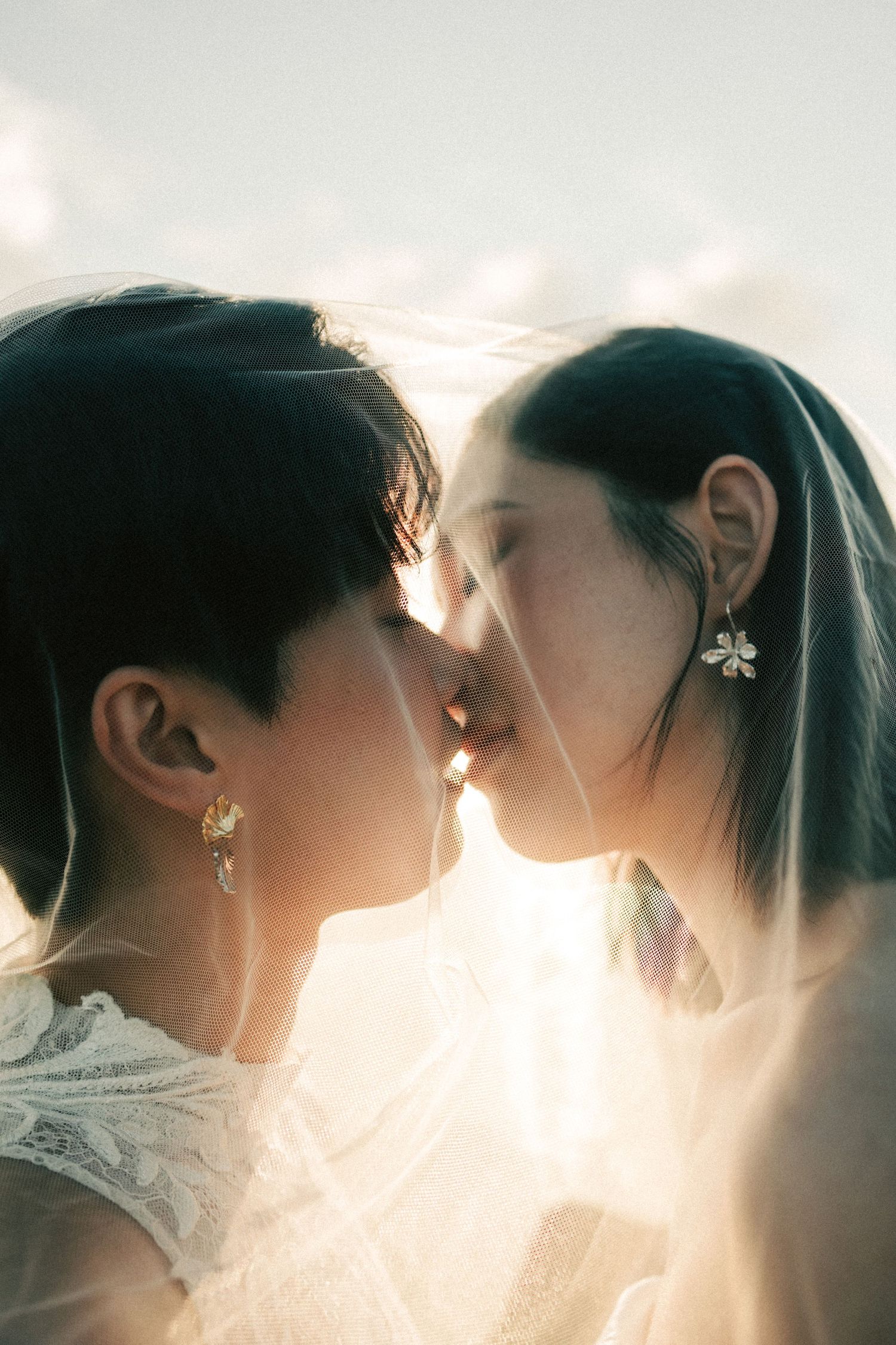 Two brides share a tender kiss under a flowing veil in soft golden sunlight.