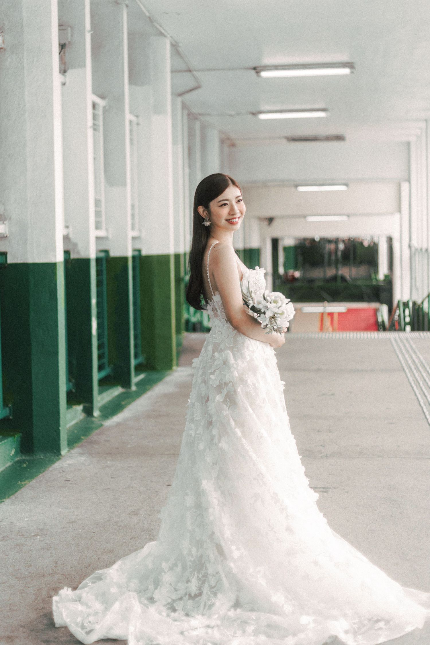 Beautiful Asian bride in a floral white wedding gown holding a bouquet in a vintage corridor.
