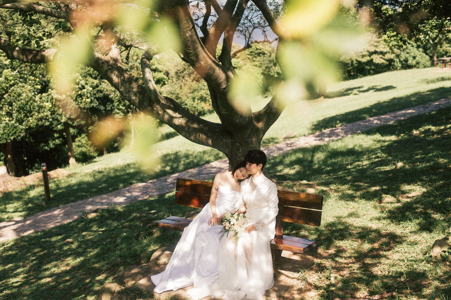 Two brides share an intimate moment seated on a wooden bench under a sprawling tree in a lush green park.