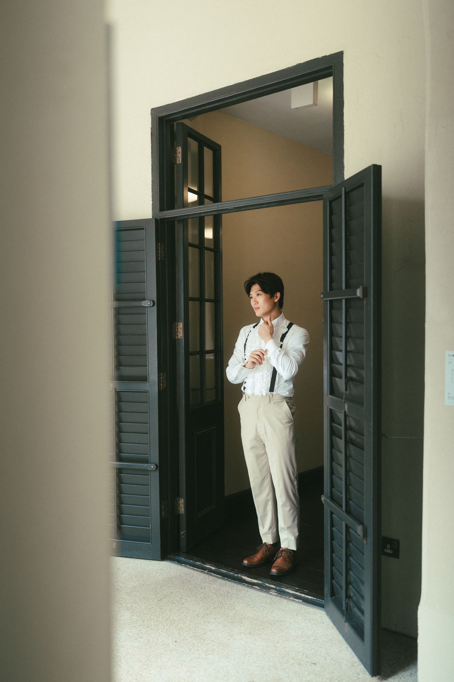 Elegant Asian man in white shirt and suspenders standing thoughtfully in a doorway.