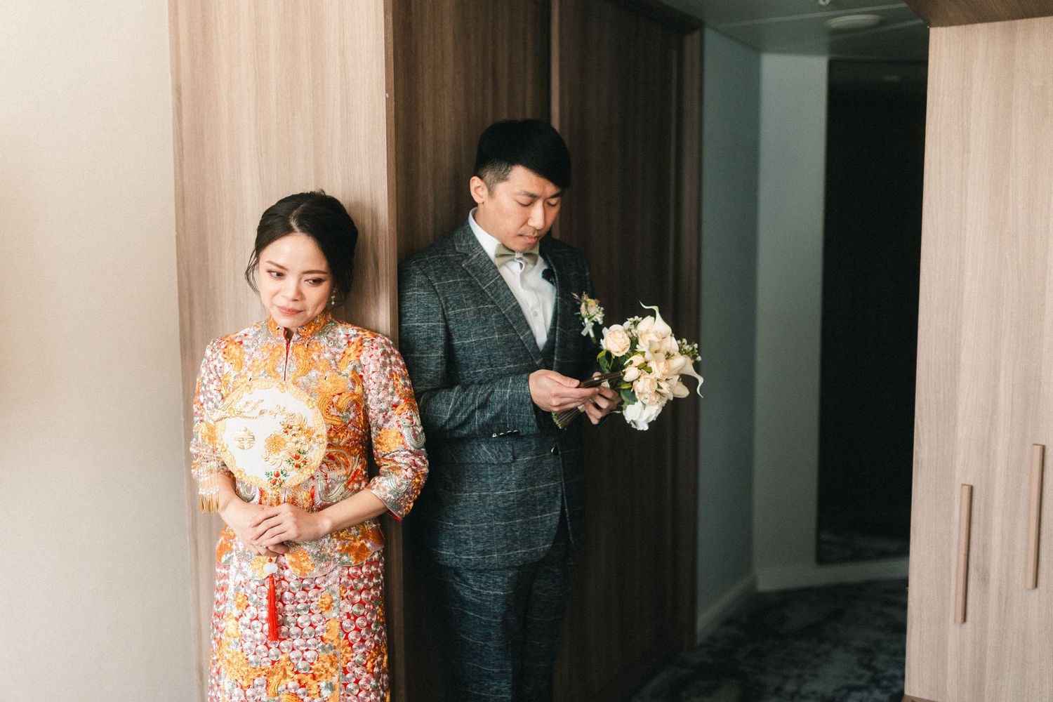 Chinese bride in ornate orange qipao holding fan stands with groom in grey suit holding white bouquet.