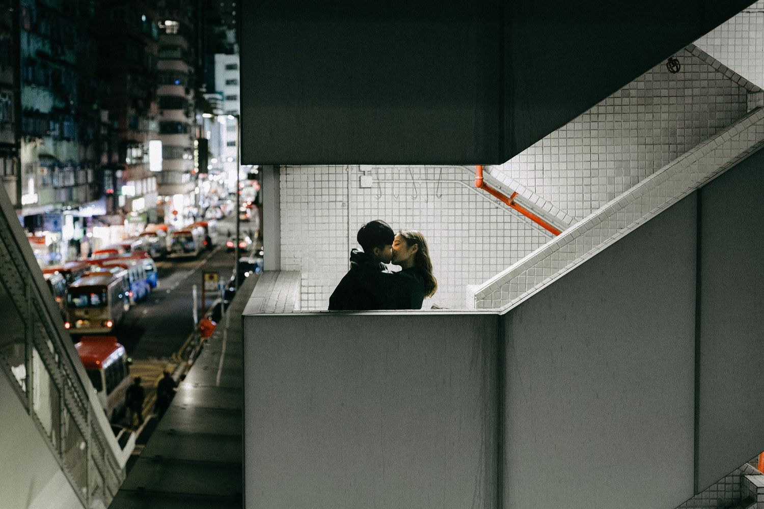A couple sharing a romantic embrace on a staircase landing with a glowing city skyline at night.