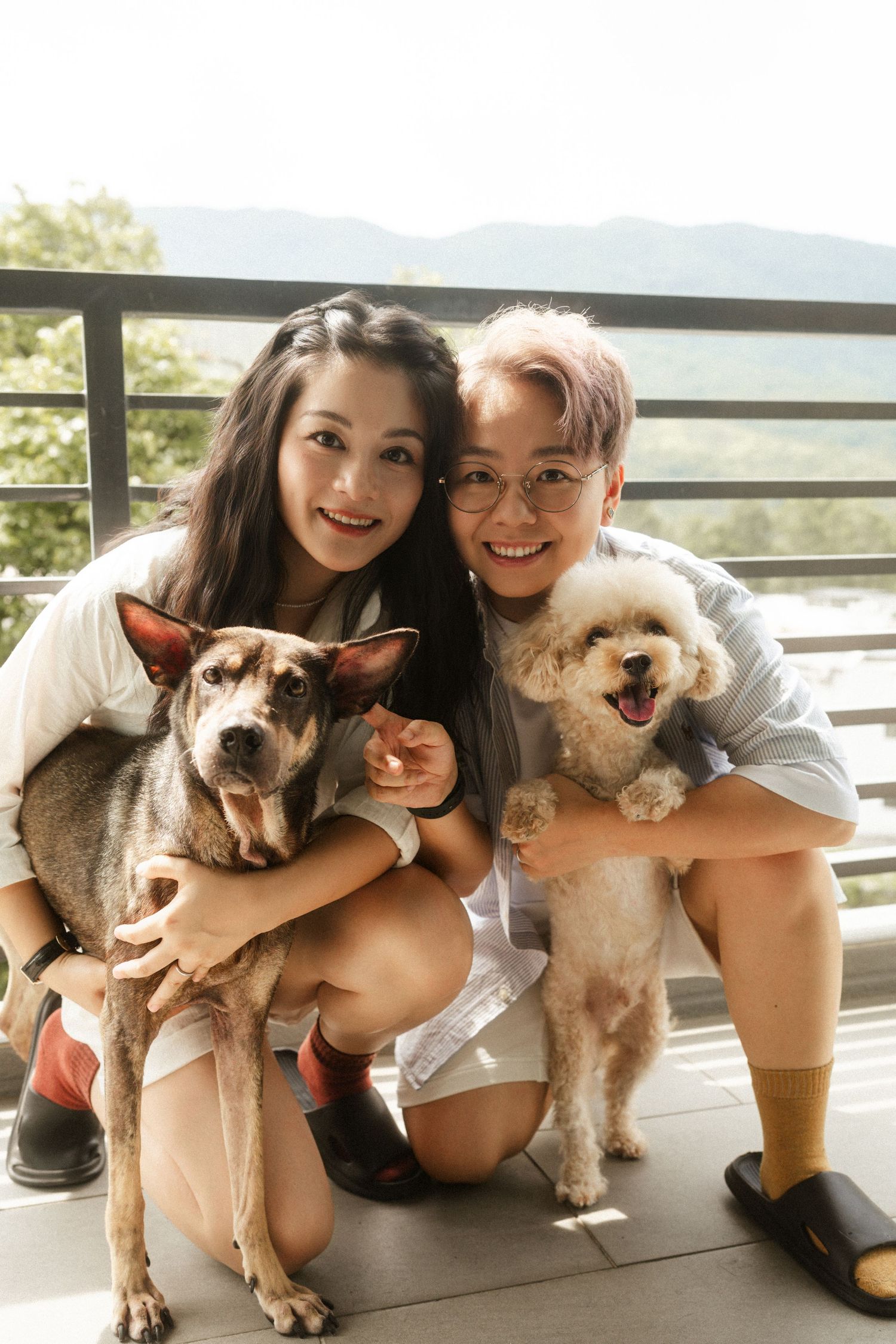 Smiling couple crouching on balcony holding two dogs, a mixed breed and a poodle, with mountains in background.
