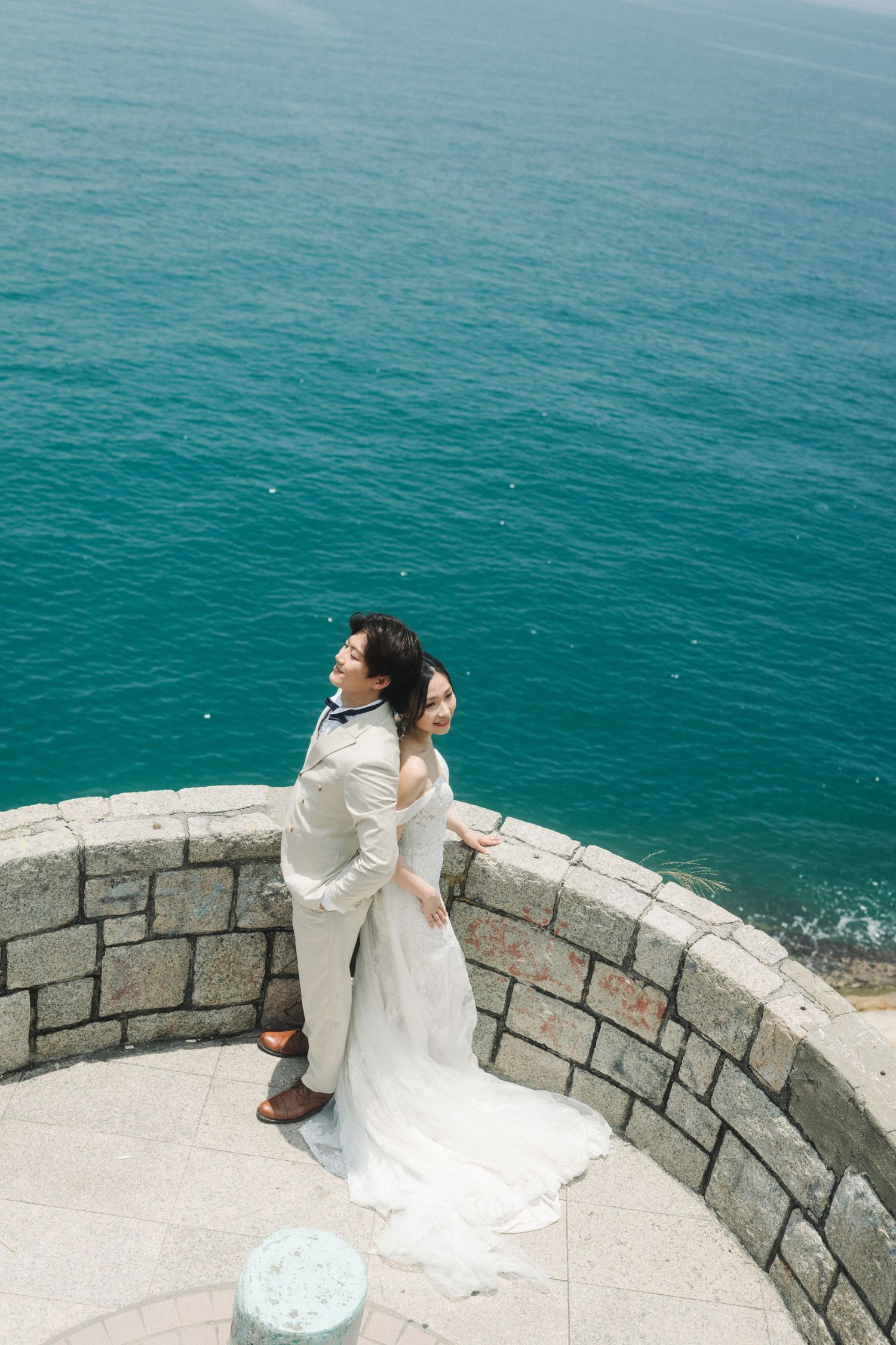 Wedding couple embrace on stone terrace overlooking stunning turquoise ocean, bride in flowing white gown.