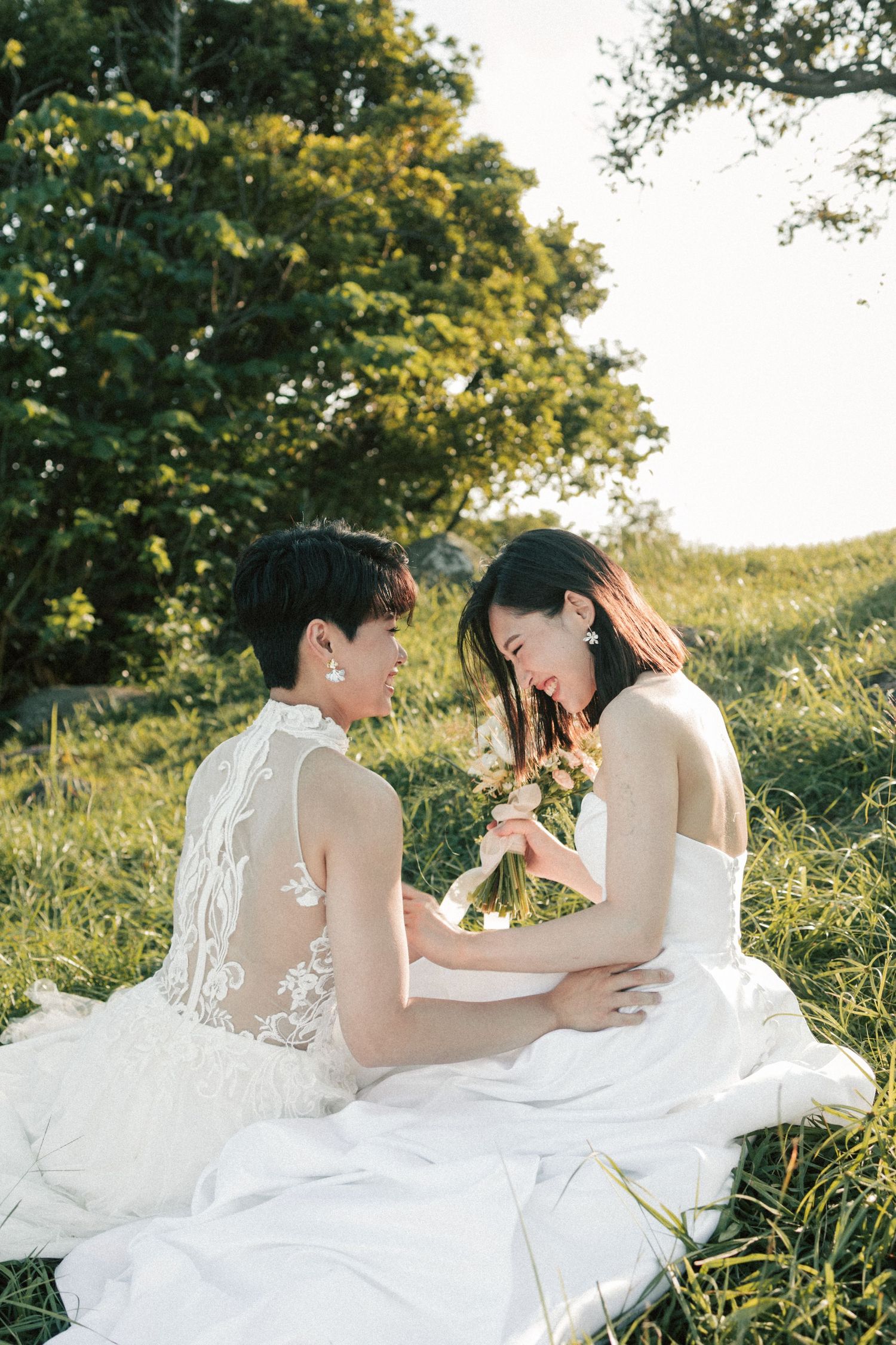 Two brides in white wedding dresses sit facing each other in a sunny green meadow.
