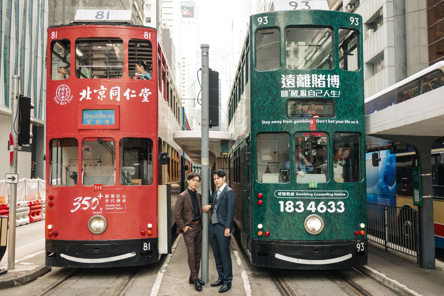 Two men in suits stand between two classic Hong Kong double-decker trams on a busy street.