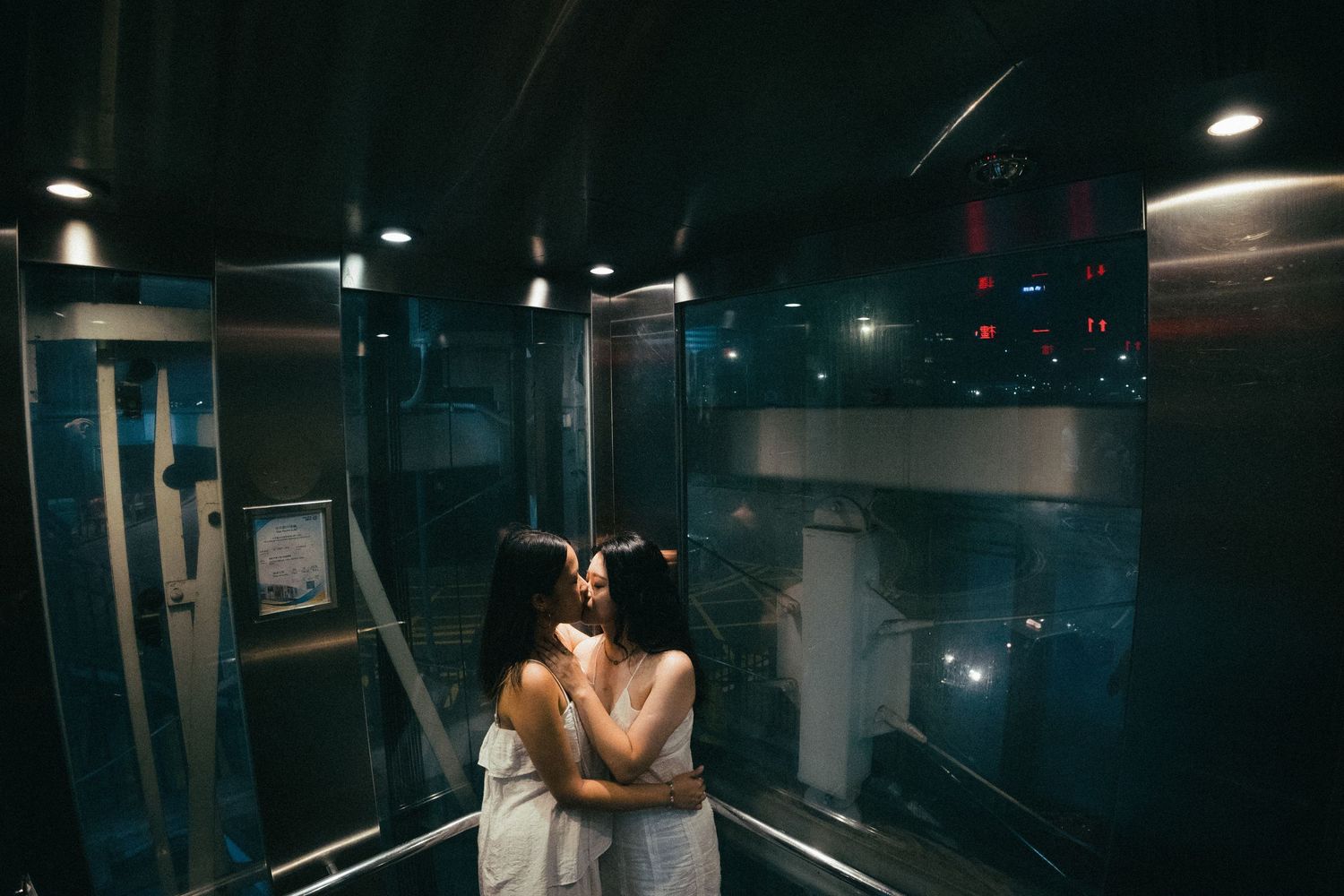 Two women in white dresses share a kiss inside a dimly lit elevator.