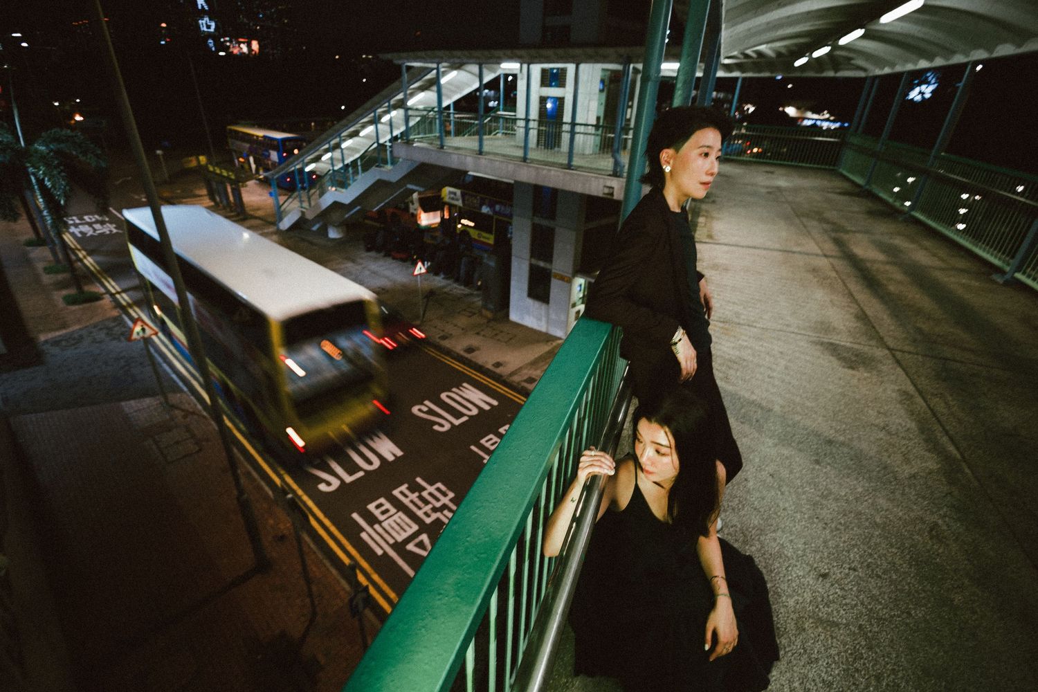 Two people dressed in black stand on an urban overpass at night with city traffic below.