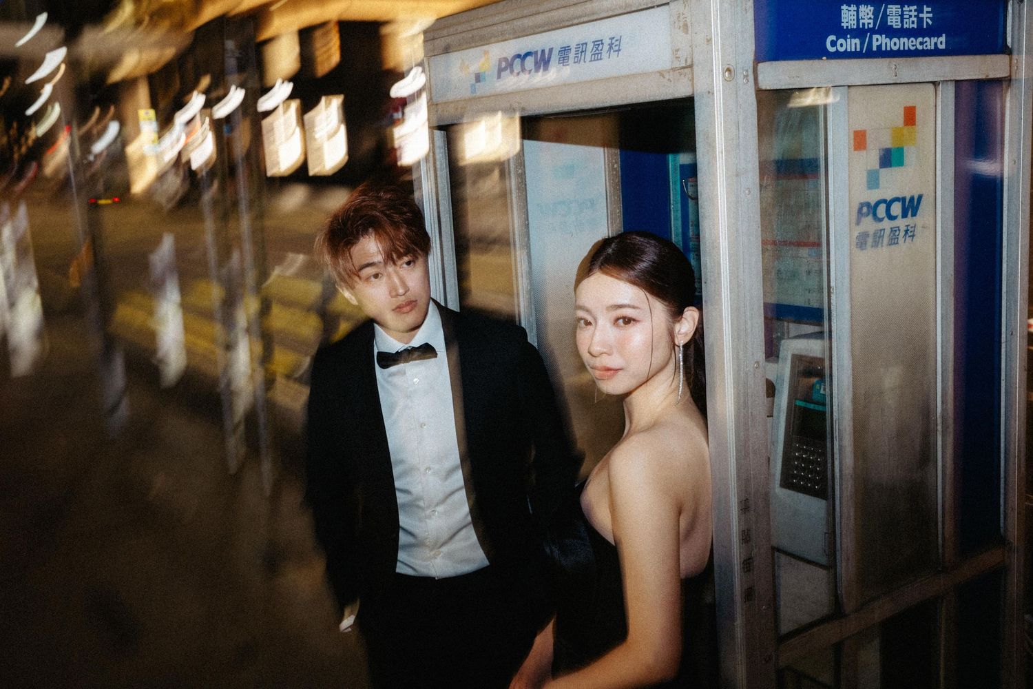 A stylish couple in black evening wear poses together by a PCCW phone booth at night in the city.