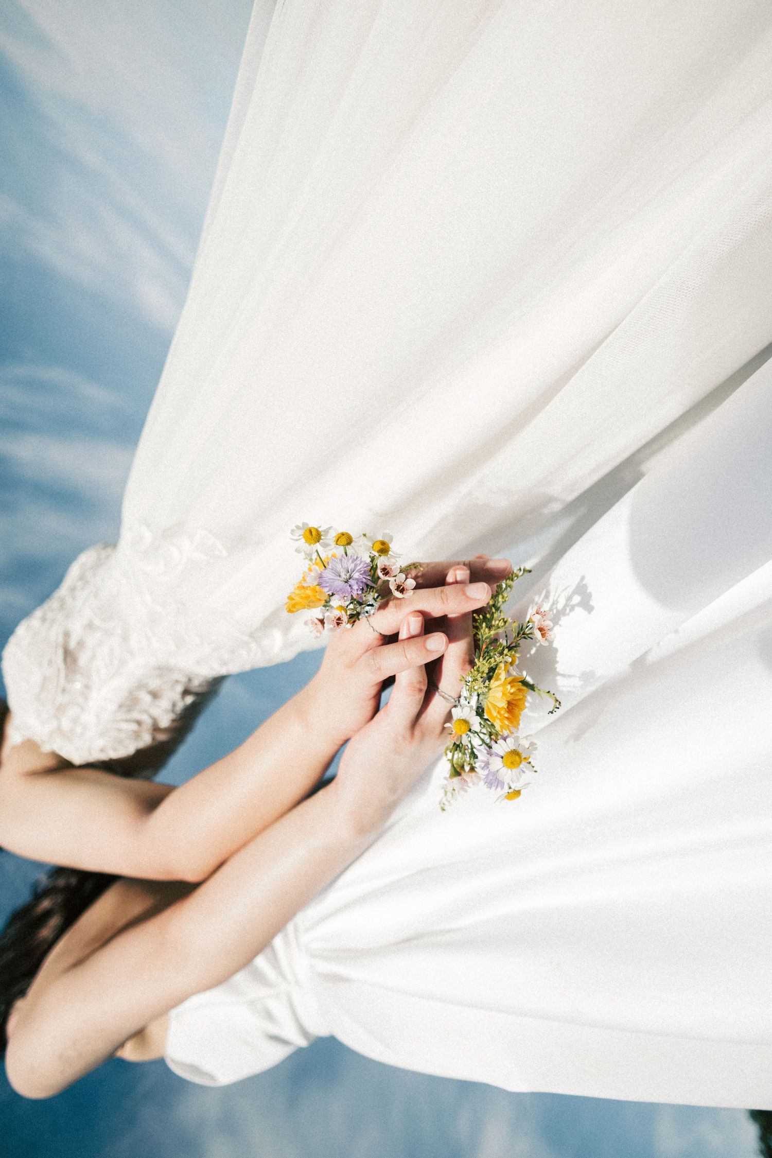Two hands adorned with wildflowers including daisies and yellow blooms rest against a flowing white wedding dress.