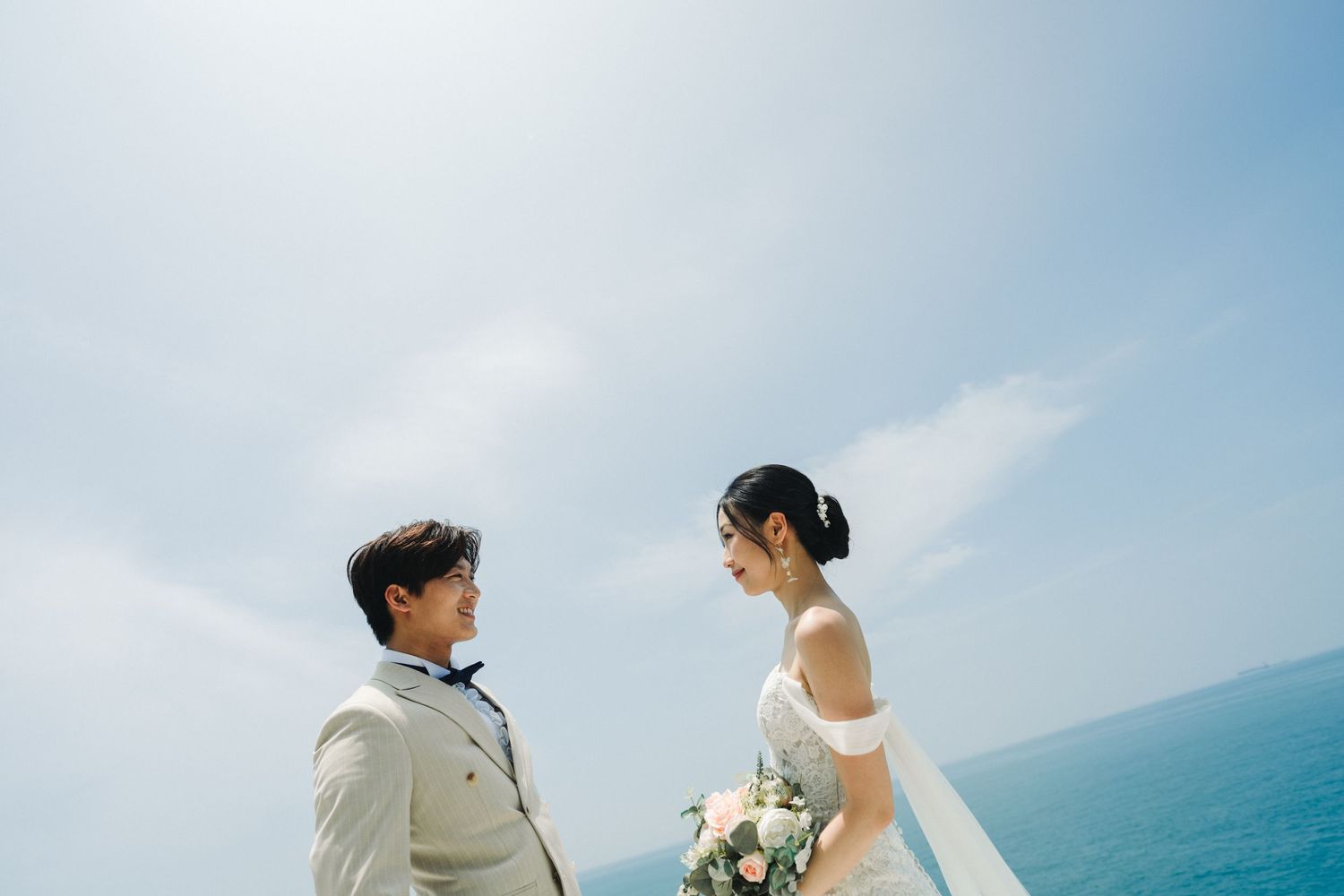 Asian wedding couple face each other against bright blue sky and turquoise ocean, bride holding bouquet.
