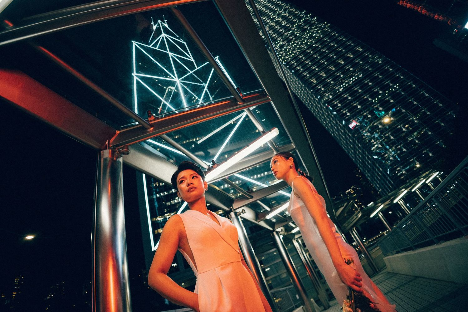 Two women in elegant outfits pose dramatically at night with illuminated Hong Kong skyscrapers in the background.