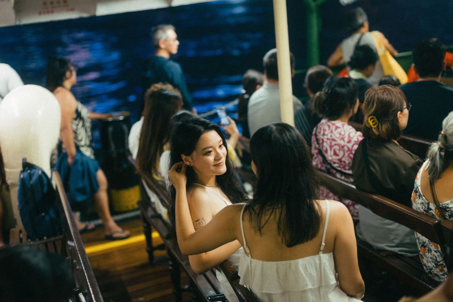 Smiling young woman in white dress sits on a ferry surrounded by other passengers at night.