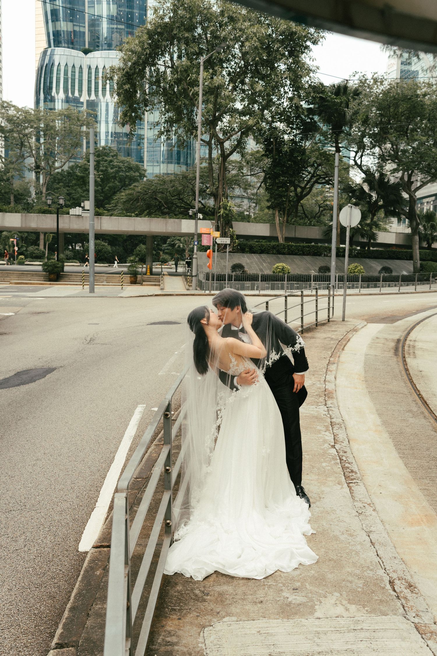 A bride and groom share a romantic kiss on a city street corner, her long veil trailing gracefully.