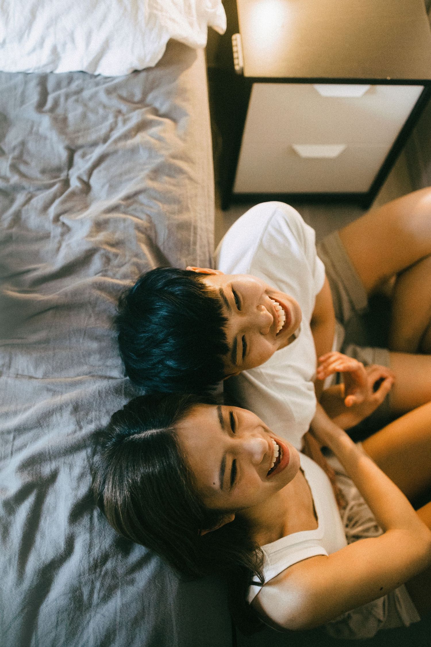 Overhead view of a smiling Asian couple lying together on a bed, laughing joyfully.