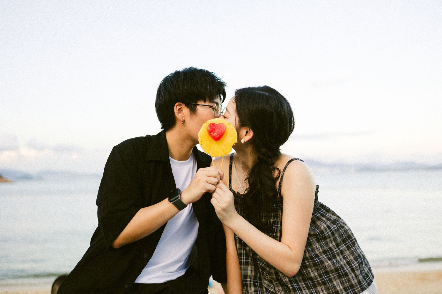 Young couple kissing behind a heart-shaped lollipop on a scenic beach at sunset.