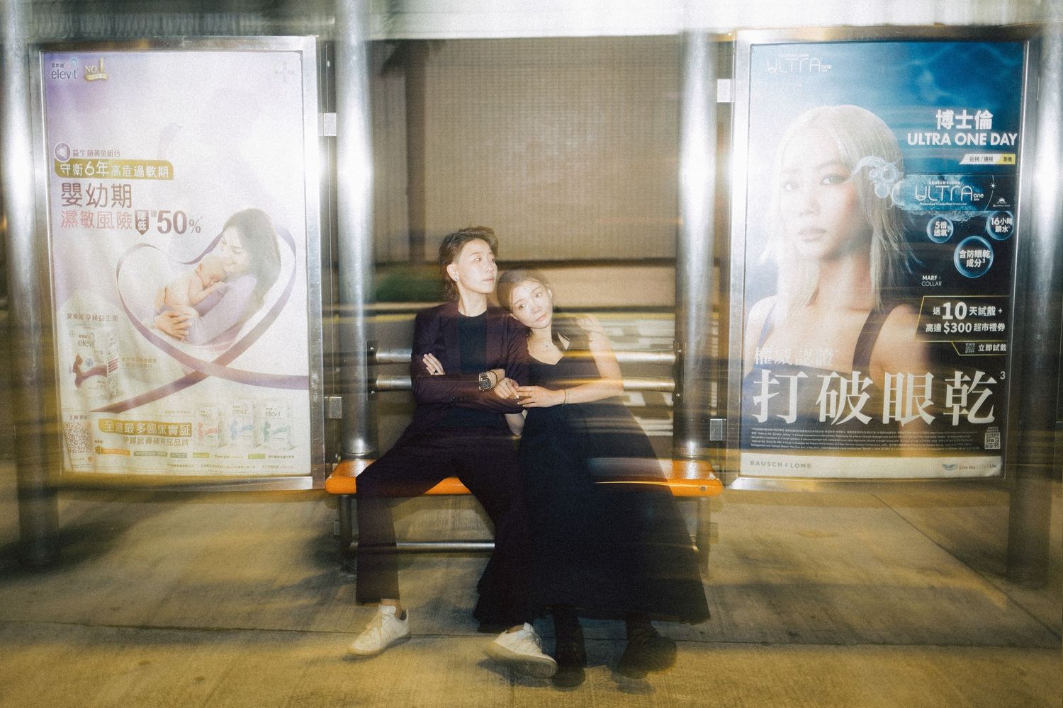 Couple sitting on a bench at a bus stop surrounded by colorful advertisement billboards.