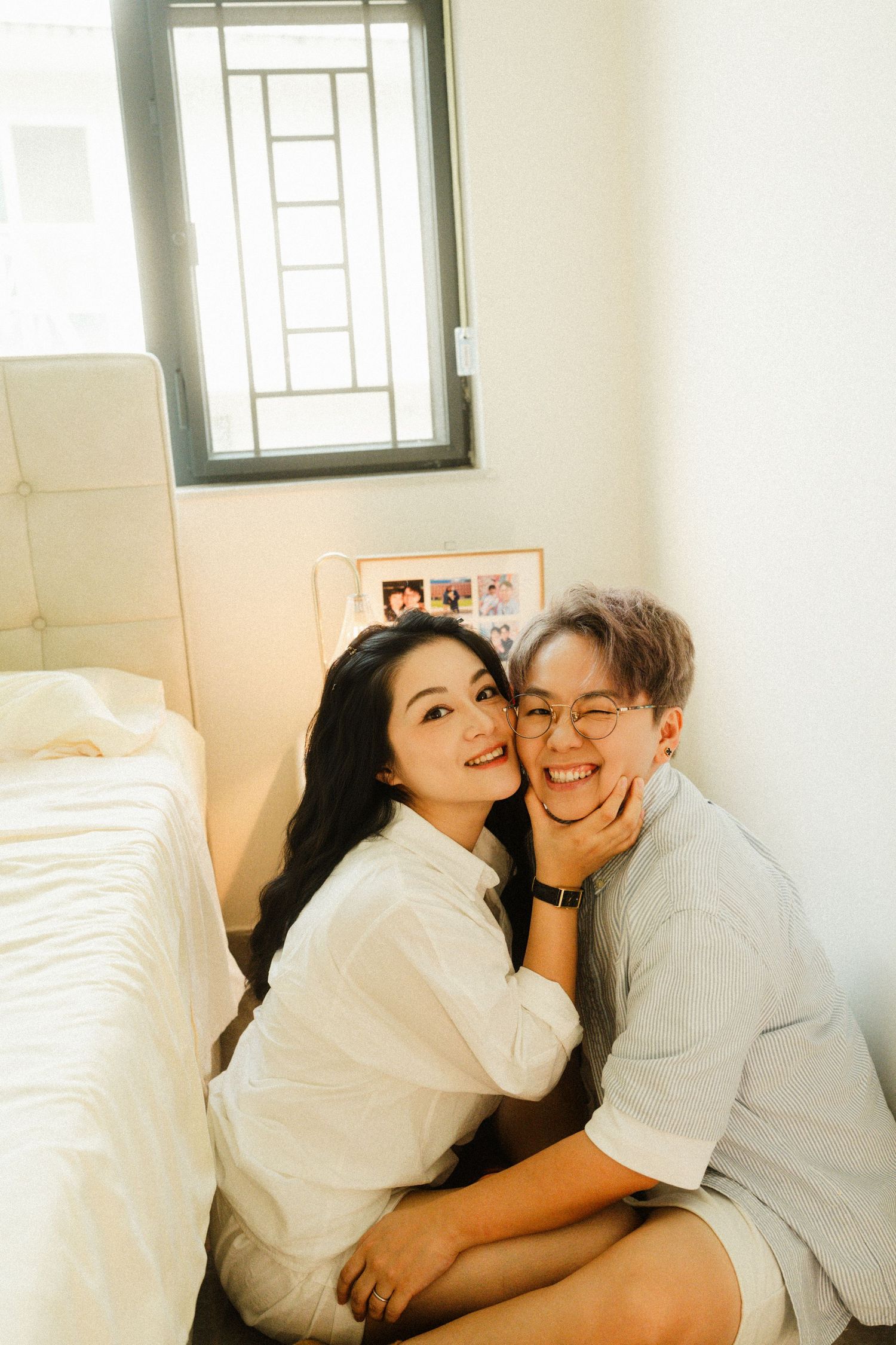Two smiling women cuddle on the floor beside a bed in a bright, cozy bedroom.